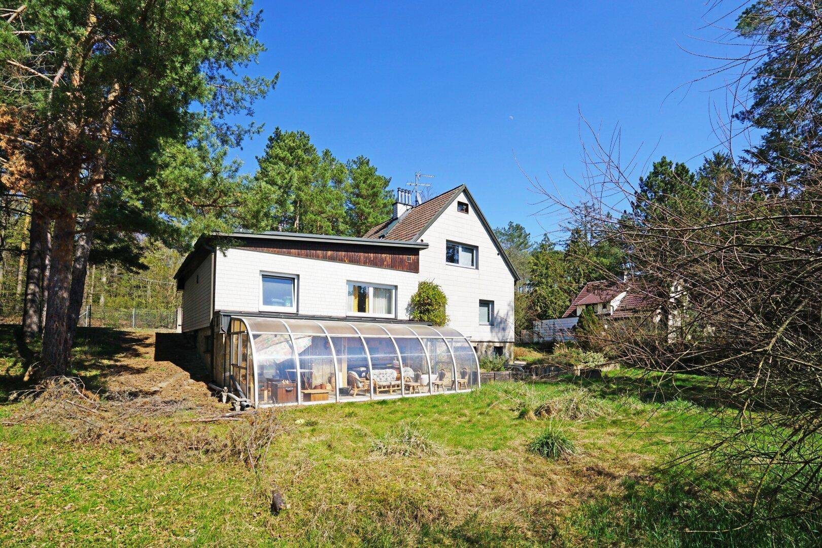Rückseite des Hauses mit einem Wintergarten, umgeben von einer weitläufigen Rasenfläche und Bäumen.