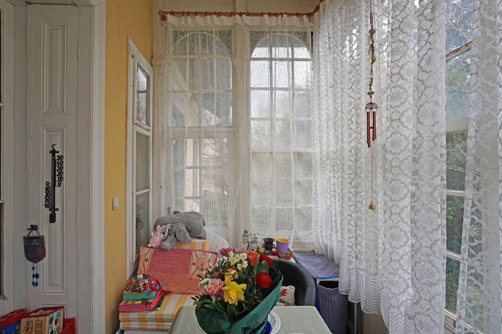 Helle Veranda mit vielen Fenstern, Vorhängen und einem kleinen Tisch mit Blumen.