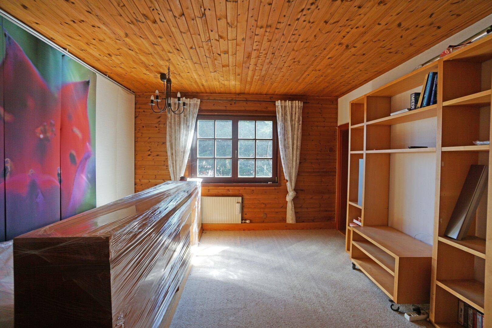 Zimmer mit Holzdecke und -wänden, Teppichboden und einem großen Fenster für Tageslicht.