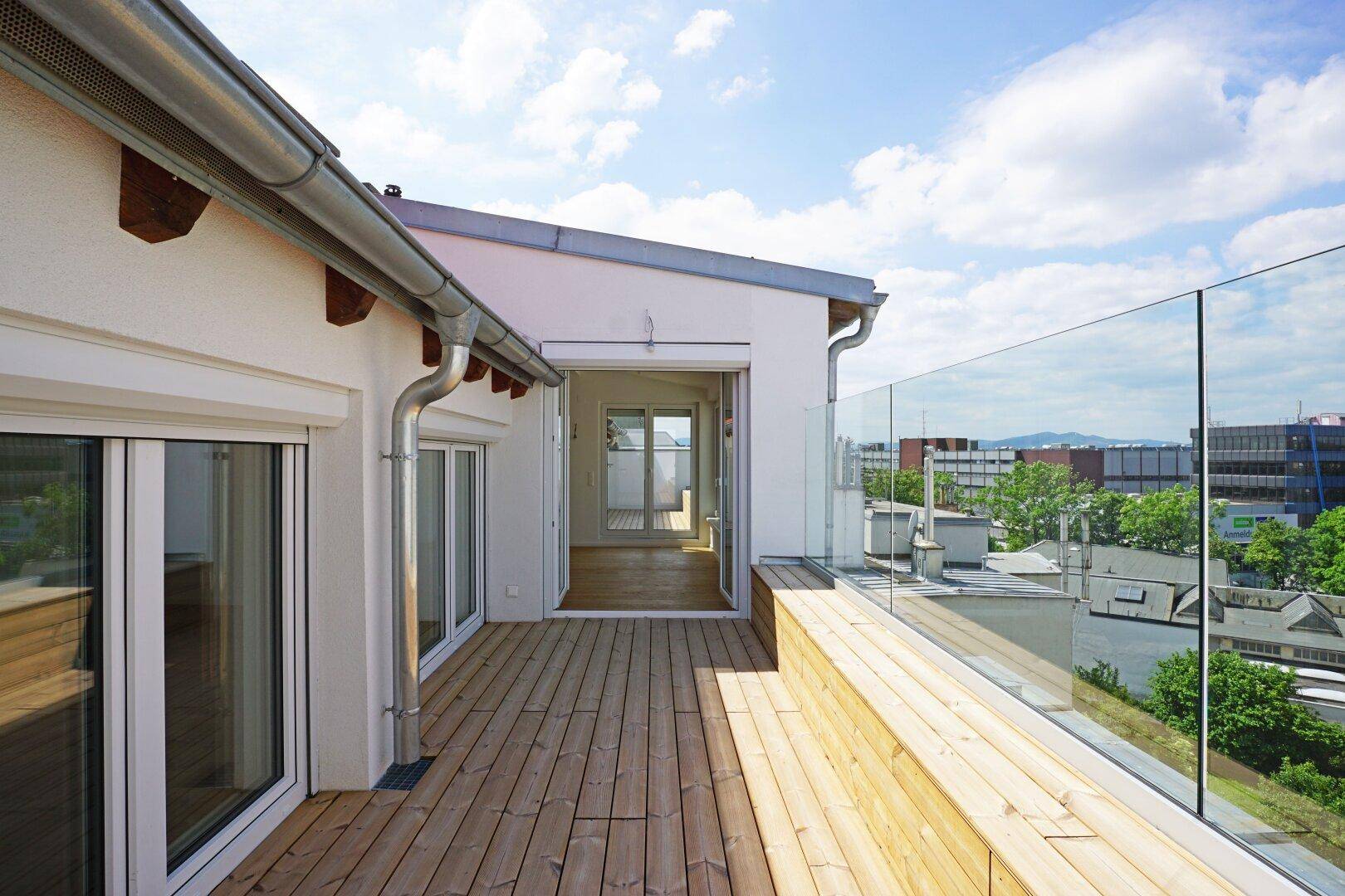Moderne Dachterrasse mit Holzplanken und Glasbrüstung, die einen weiten Blick über die Stadt ermöglicht.