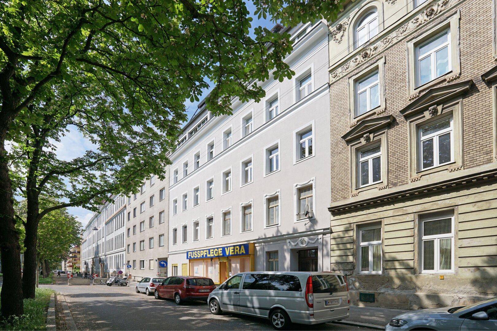 Historisches Gebäude mit klassischer Fassade an einer belebten Straße mit Bäumen und parkenden Autos.
