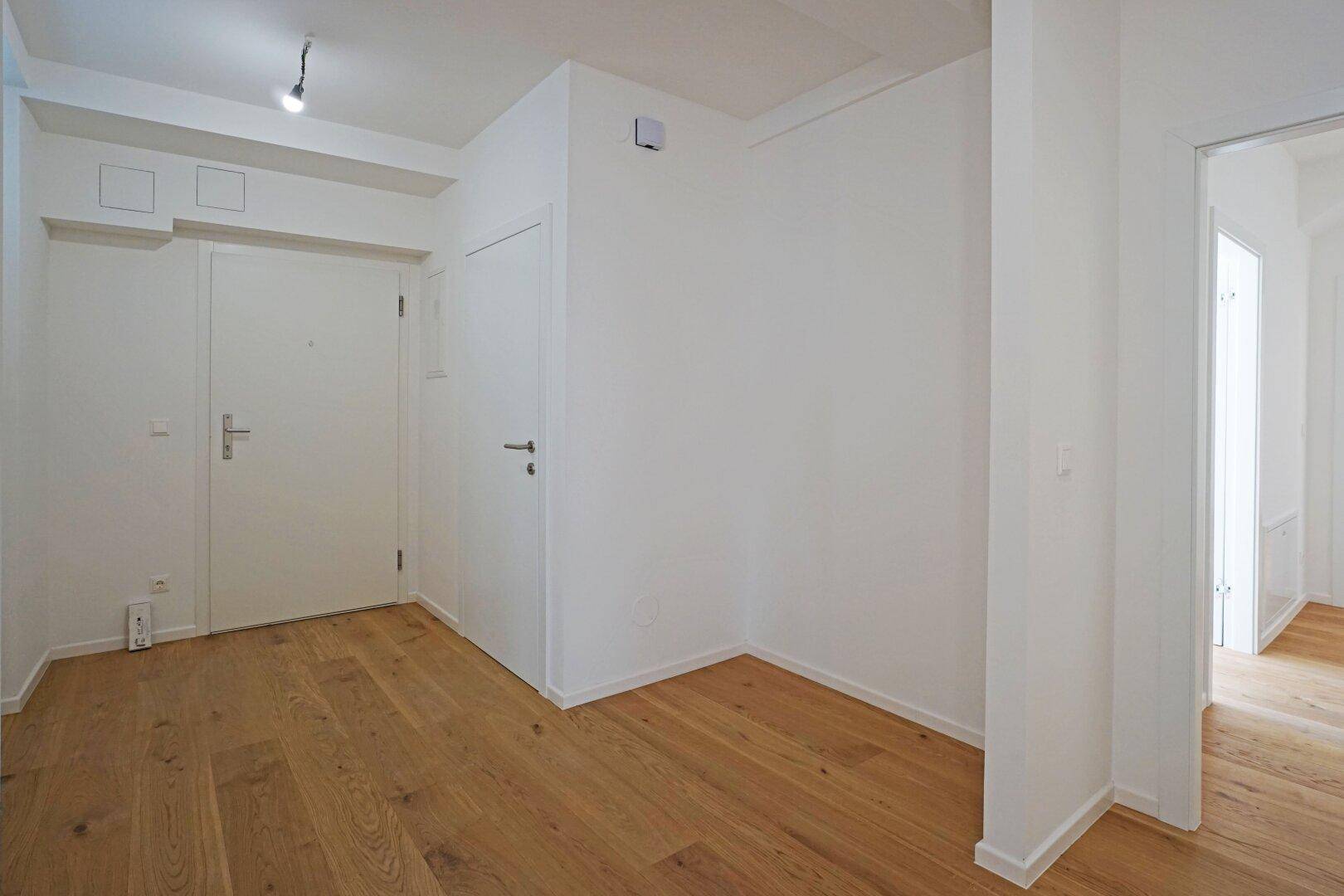 Heller Eingangsbereich mit Holzboden und weißen Wänden, der zu weiteren Räumen führt.