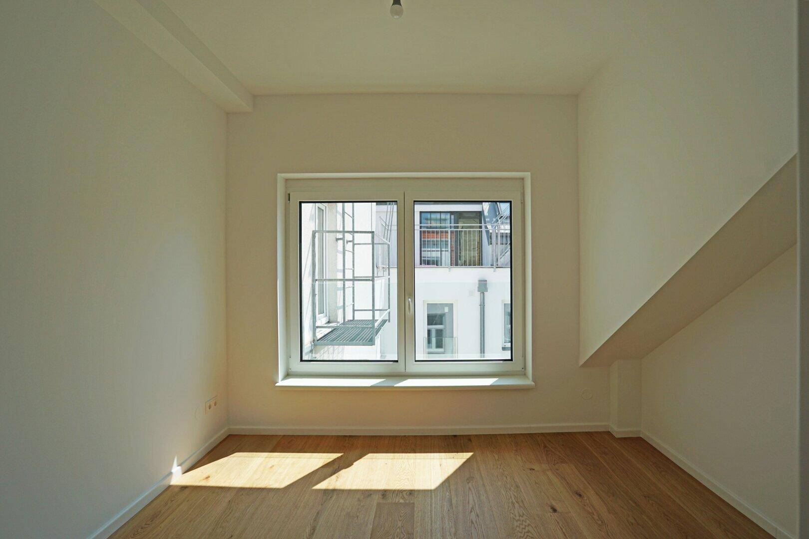 Freundliches Zimmer mit Holzboden und einem großen Fenster, das viel Tageslicht hereinlässt.