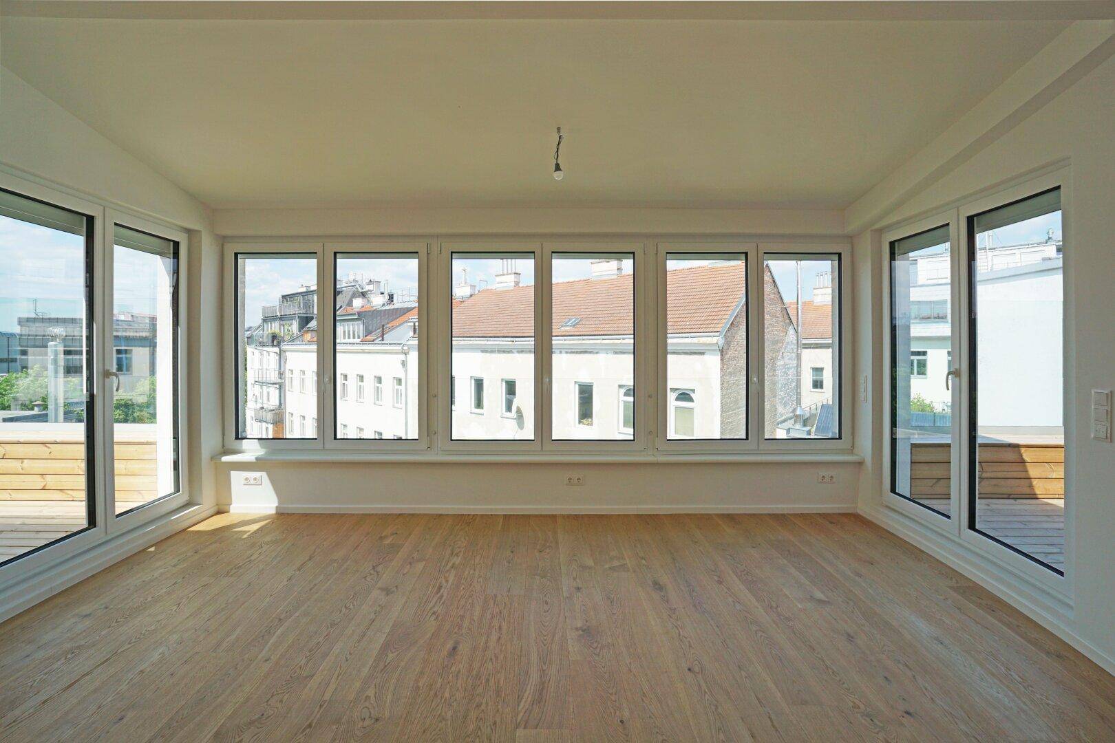 Helles, leeres Zimmer mit großen Fenstern und Holzboden, bietet einen weiten Blick über die Stadt.