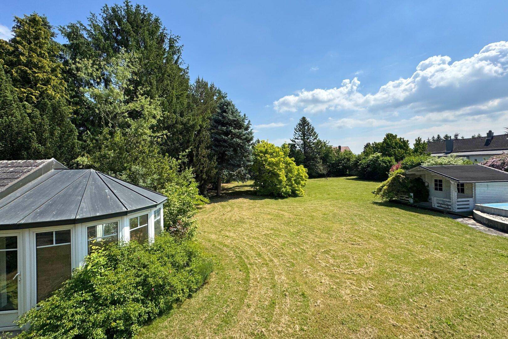 Weitläufiger Garten mit Rasenfläche, Bäumen, Gartenhaus und Pool unter blauem Himmel.