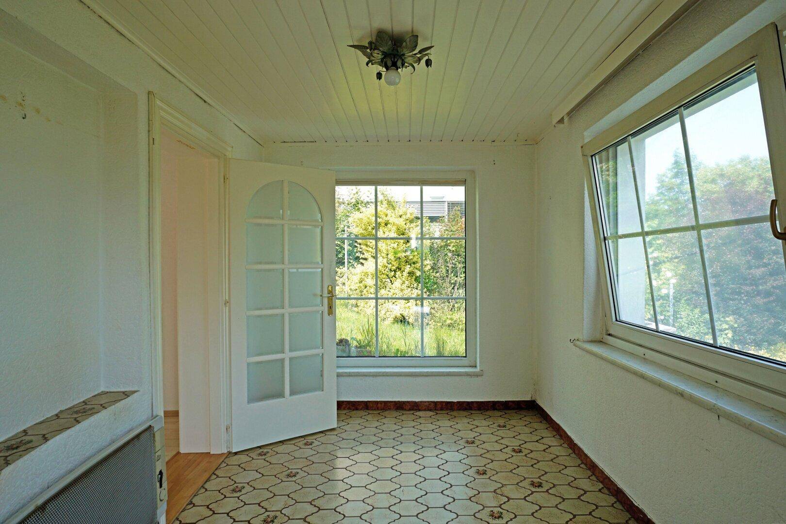 Heller Innenraum mit gemustertem Fliesenboden, Holzdecke und großem Fenster mit Blick ins Grüne.