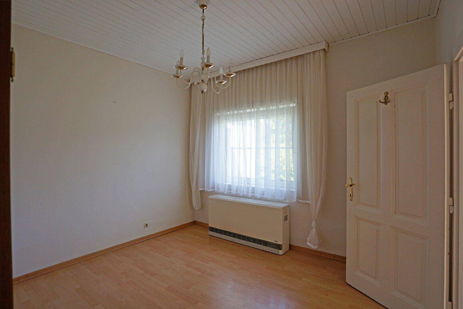 Schlafzimmer mit hellem Holzboden, Fenster mit Vorhängen und einer Holzpaneeldecke, beleuchtet von einem Kronleuchter.