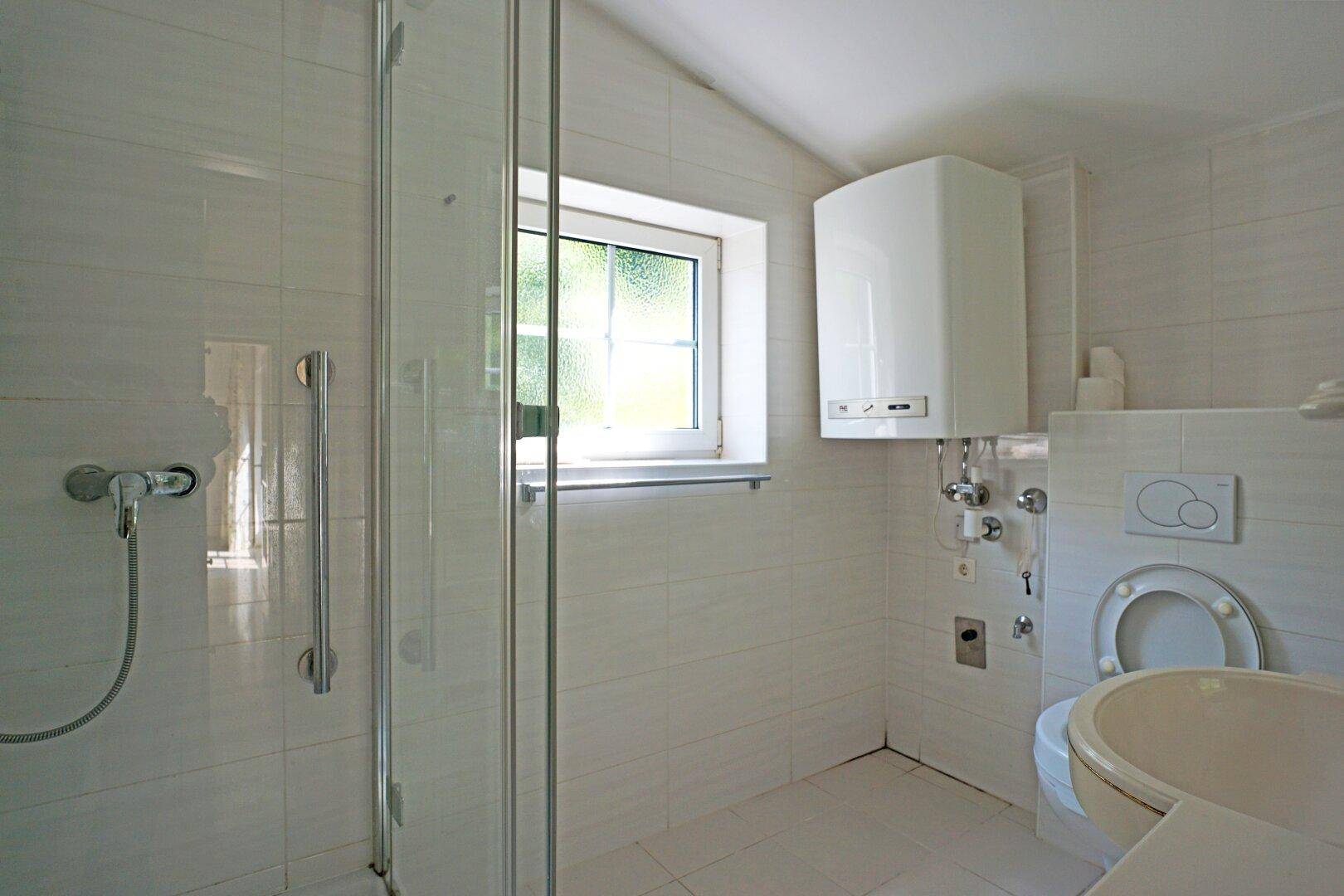 Badezimmer mit weißen Fliesen, Duschkabine, WC und Fenster, ausgestattet mit Haltegriffen für Barrierefreiheit.