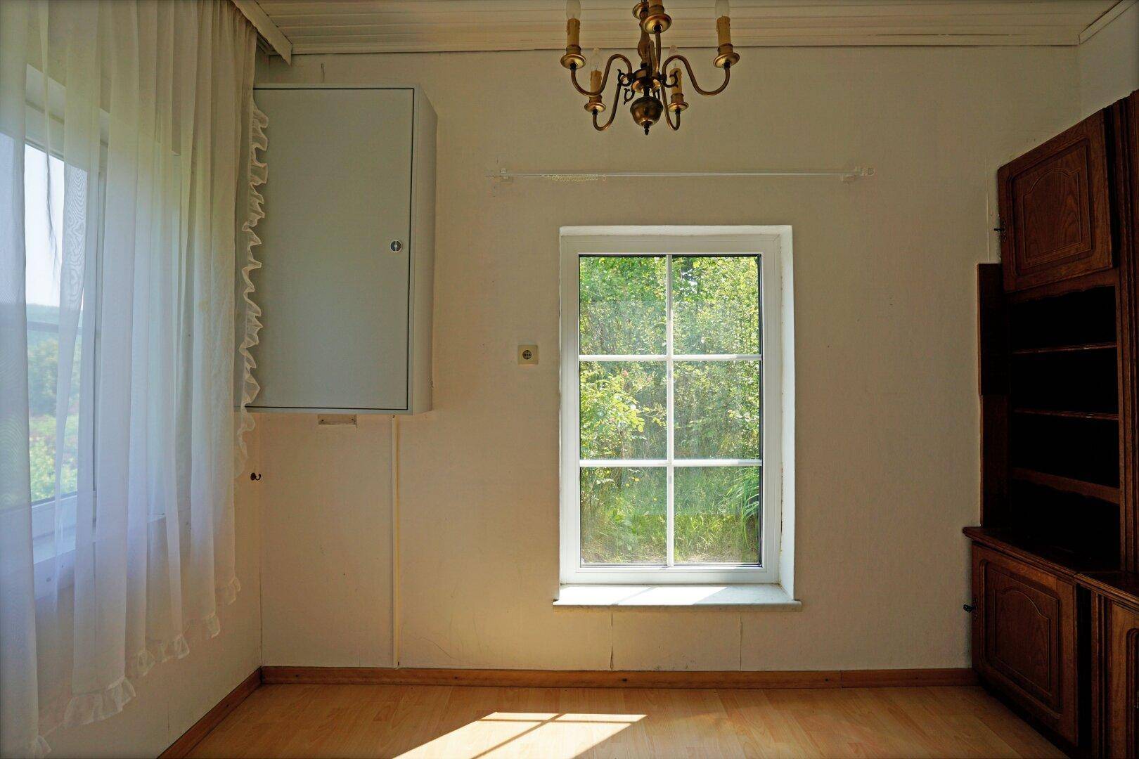 Heller Raum mit Holzboden, großem Fenster mit Blick ins Grüne und einem traditionellen Holzschrank.
