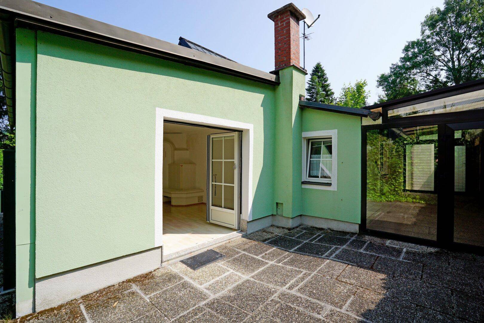 Gepflasterte Terrasse vor dem hellgrünen Haus mit offener Tür und einem angrenzenden überdachten Gang.