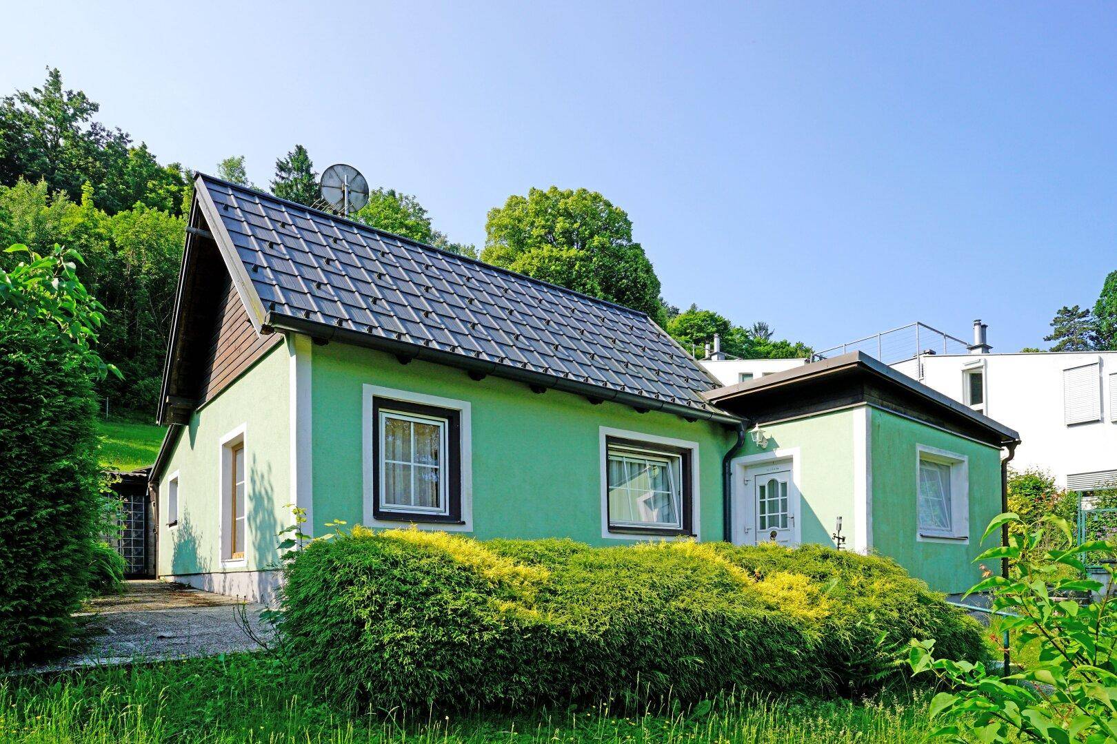 Charmantes hellgrünes Haus mit dunklem Ziegeldach und gepflegten Büschen vor den Fenstern.