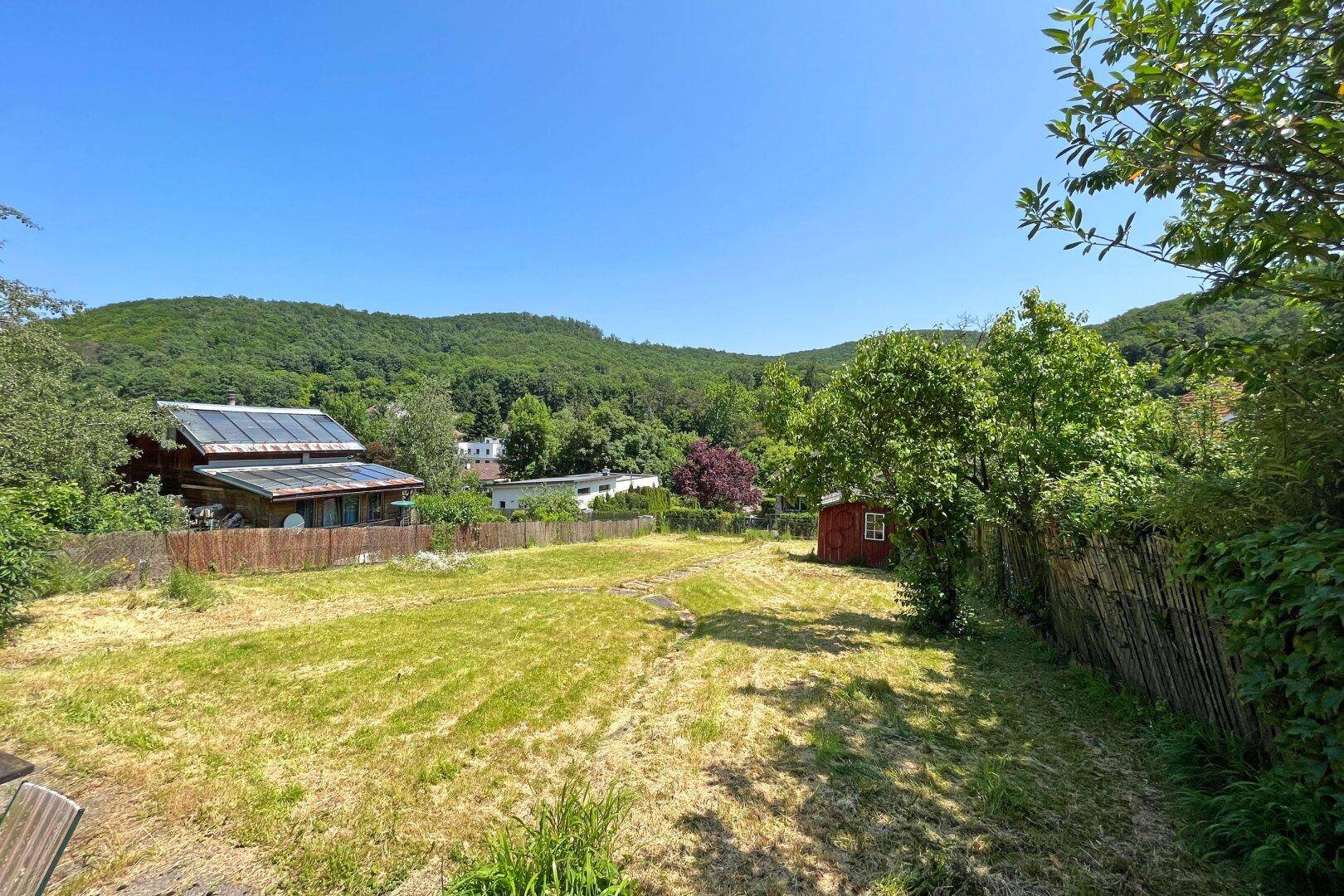 Weitläufige Gartenfläche mit einem kleinen roten Schuppen und Blick auf grüne Hügel unter blauem Himmel.