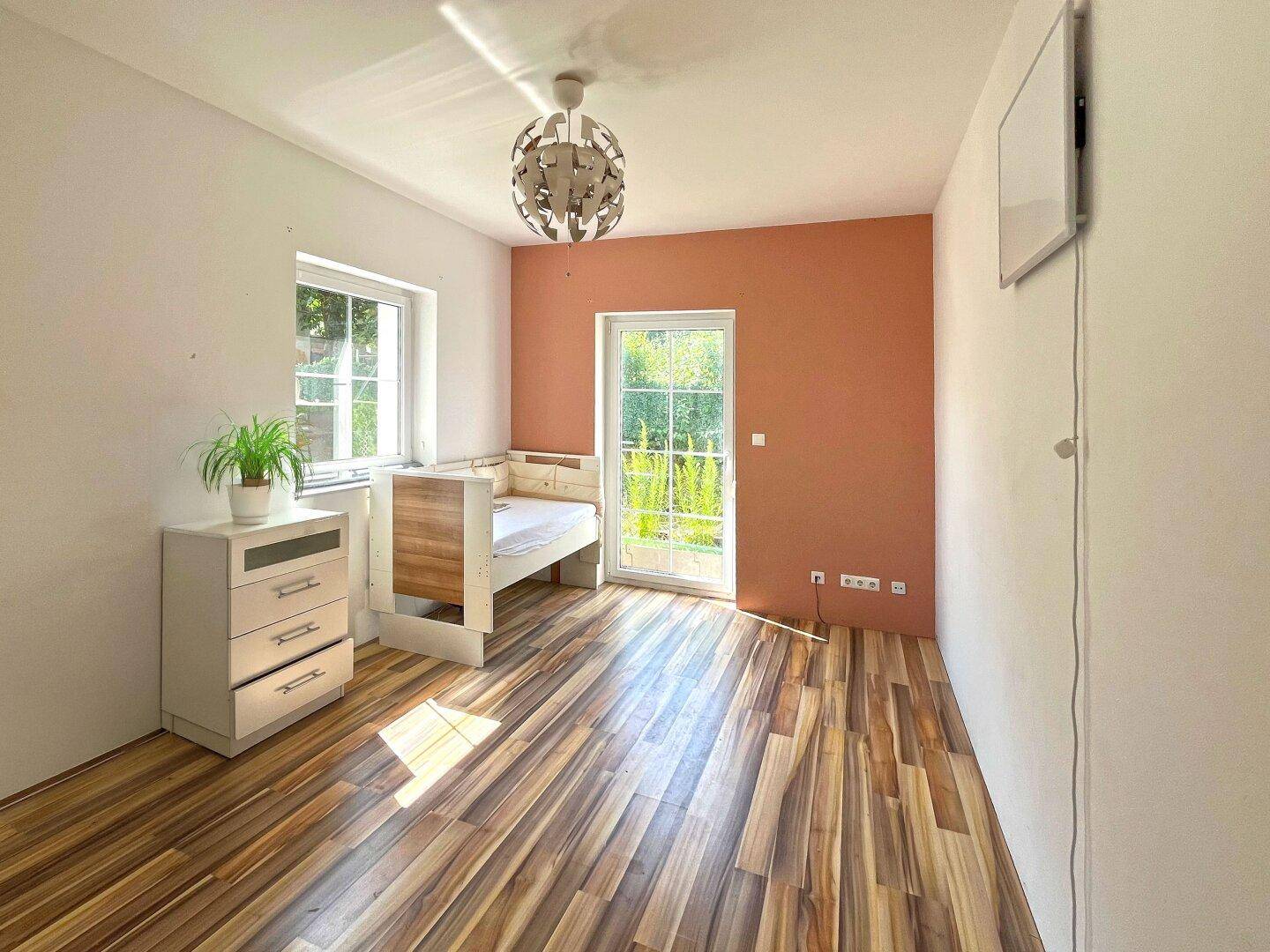 Helles Schlafzimmer mit Holzboden, farbiger Akzentwand und Zugang zum Außenbereich.