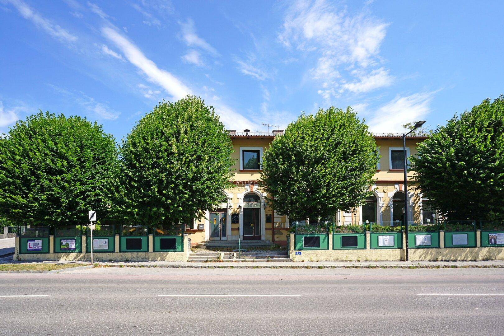 Historisches Gebäude mit gelber Fassade und grünen Bäumen entlang der Straße unter blauem Himmel.