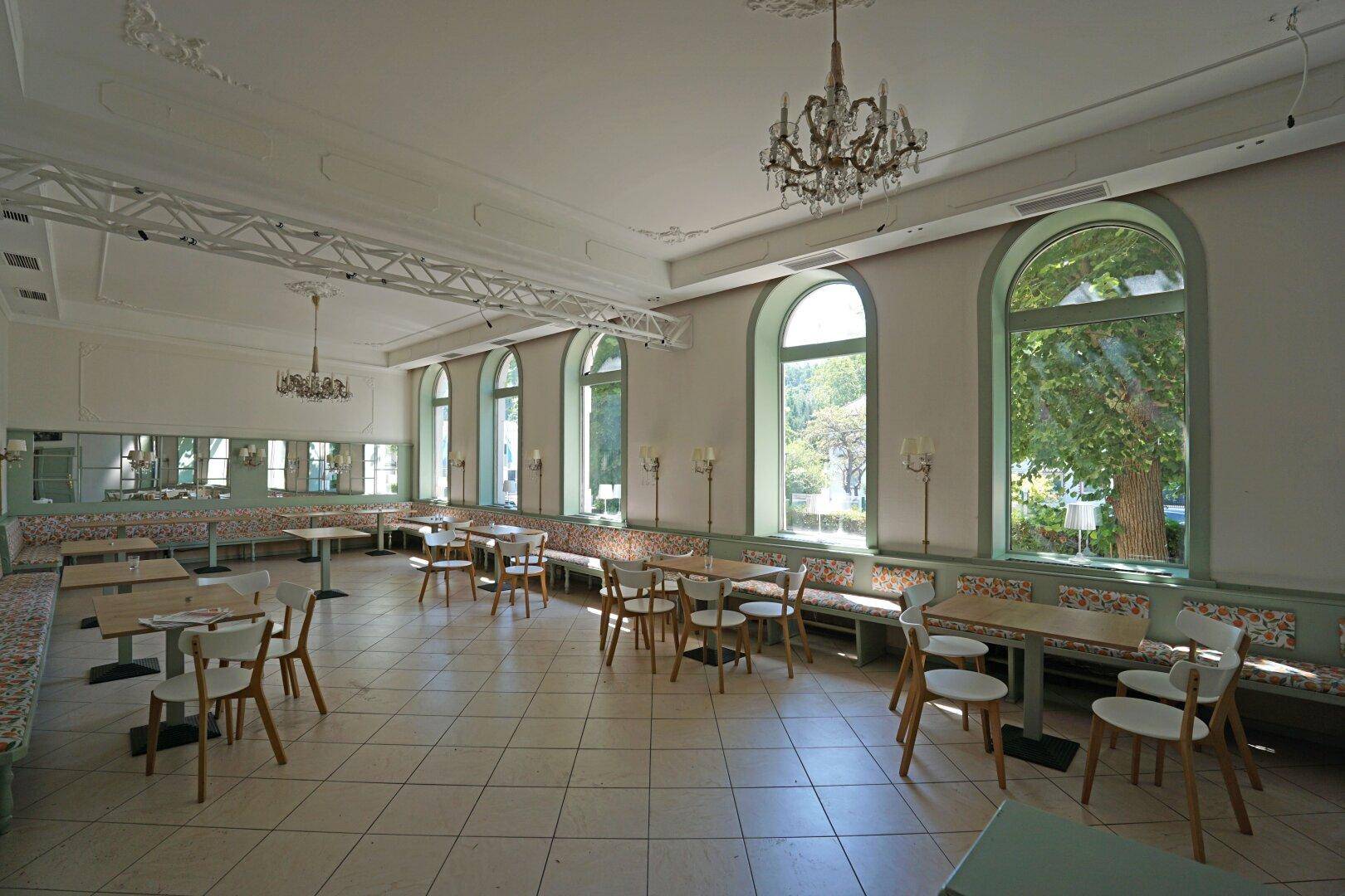 Heller Speisesaal mit Kronleuchter, gepolsterten Sitzbänken und Blick ins Grüne durch Rundbogenfenster.