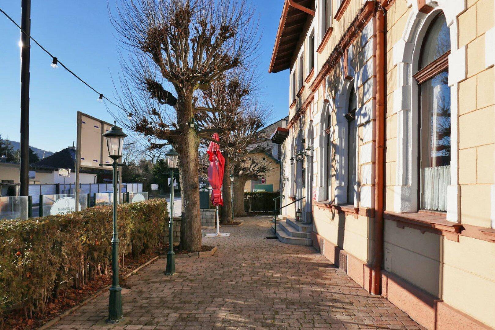 Seitenansicht eines historischen Gebäudes mit gepflastertem Weg und kahlen Bäumen im Winterlicht.