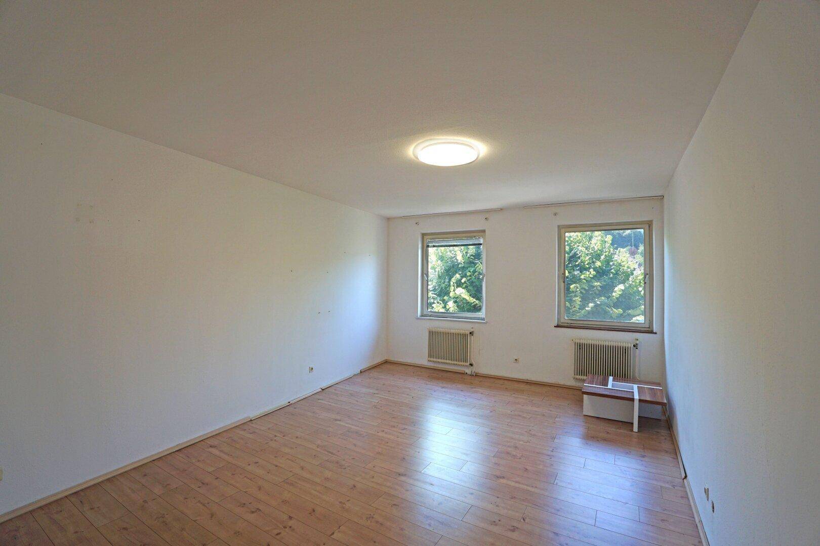 Leeres Zimmer mit Holzfußboden, zwei Fenstern und Heizkörpern unter der Decke.
