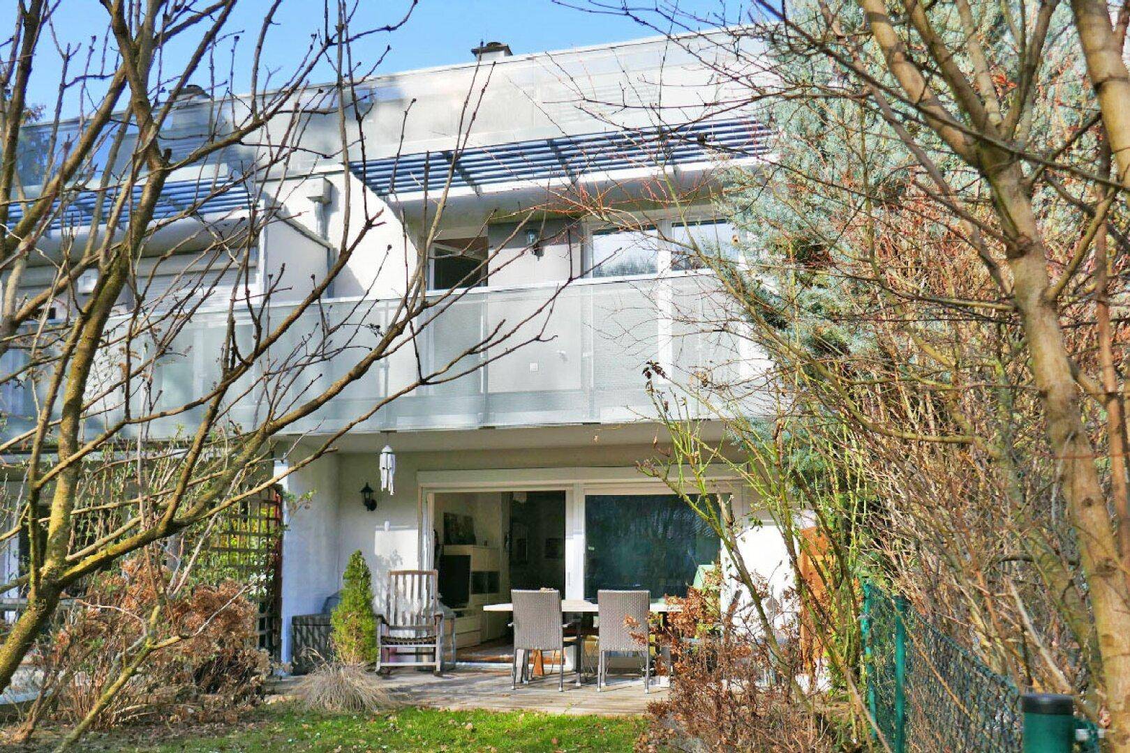 Ansicht des Hauses von hinten mit Terrasse und Balkonen in einer grünen, baumbestandenen Umgebung.