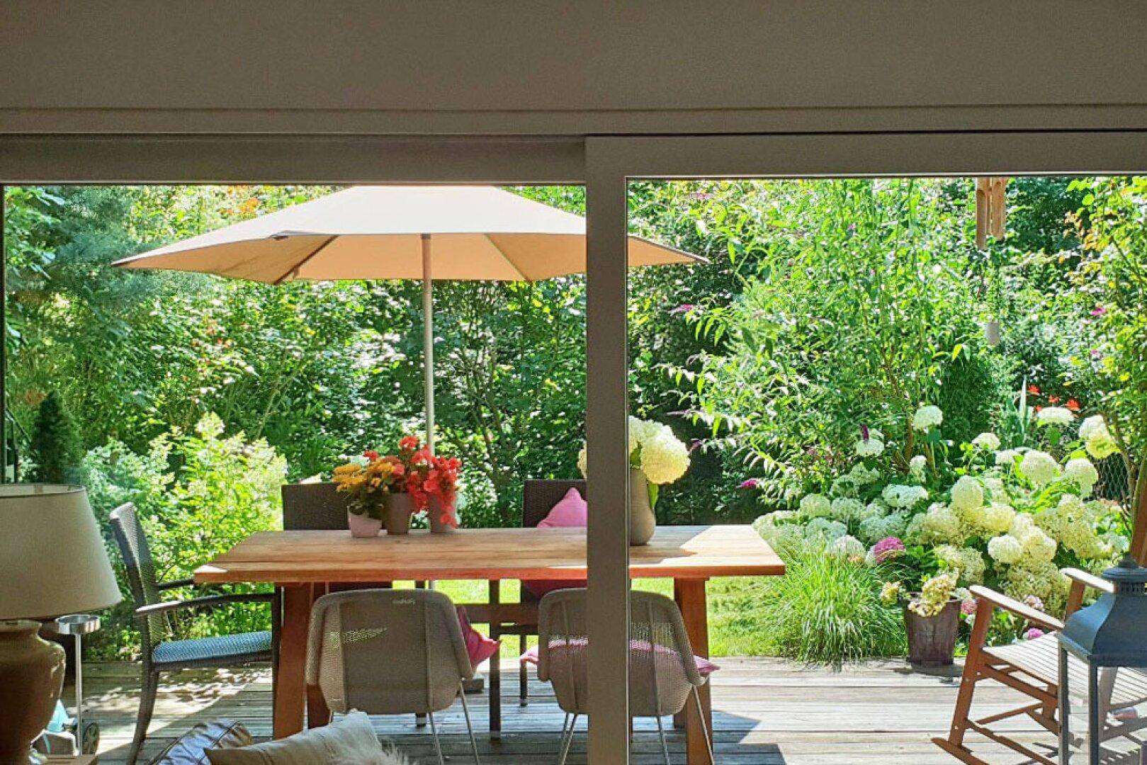 Blick vom Wohnzimmer auf die grüne Terrasse mit Esstisch, Sonnenschirm und üppiger Bepflanzung.