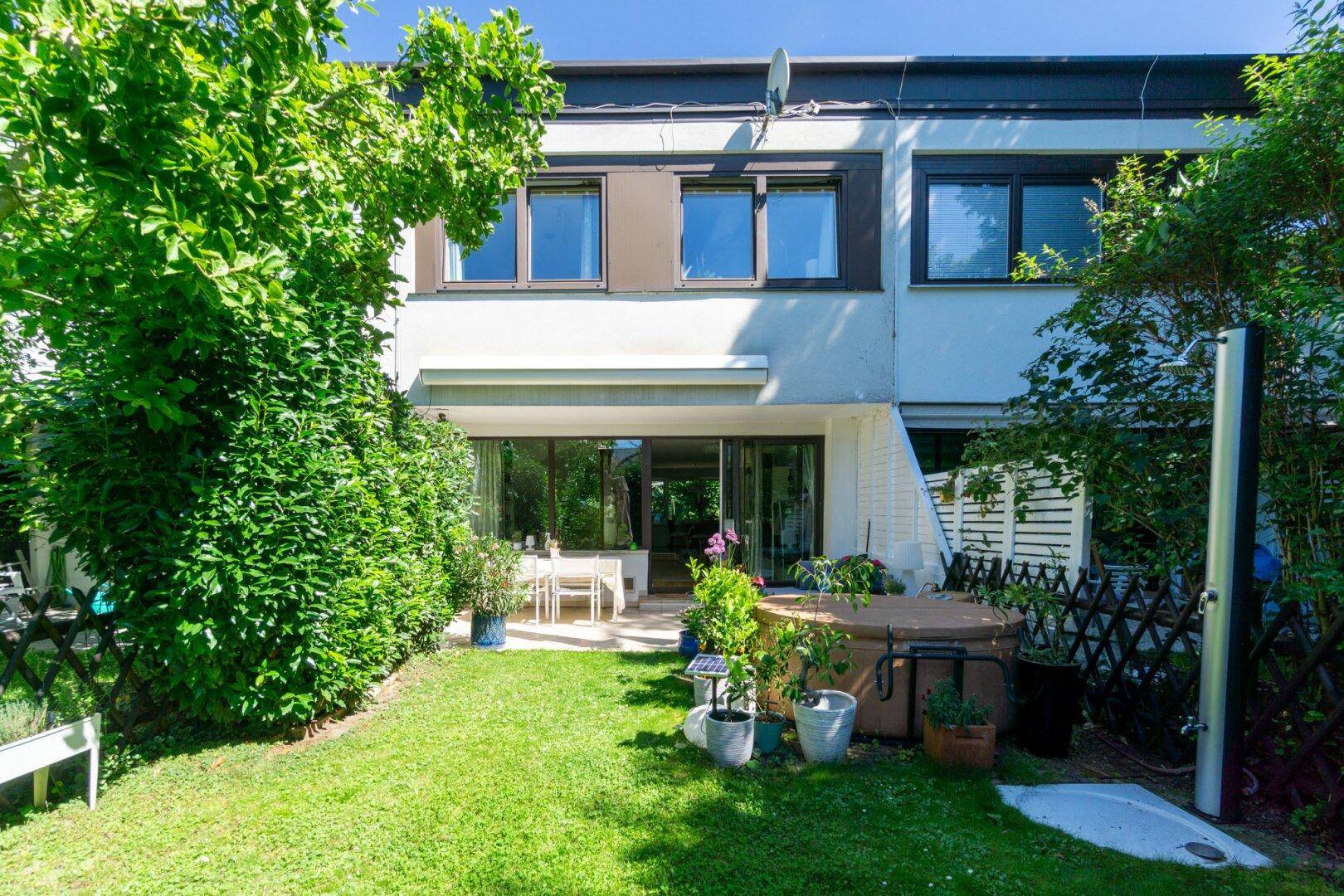 Sonniger Garten mit Terrasse, Rasen und Außendusche, umgeben von üppigem Grün.