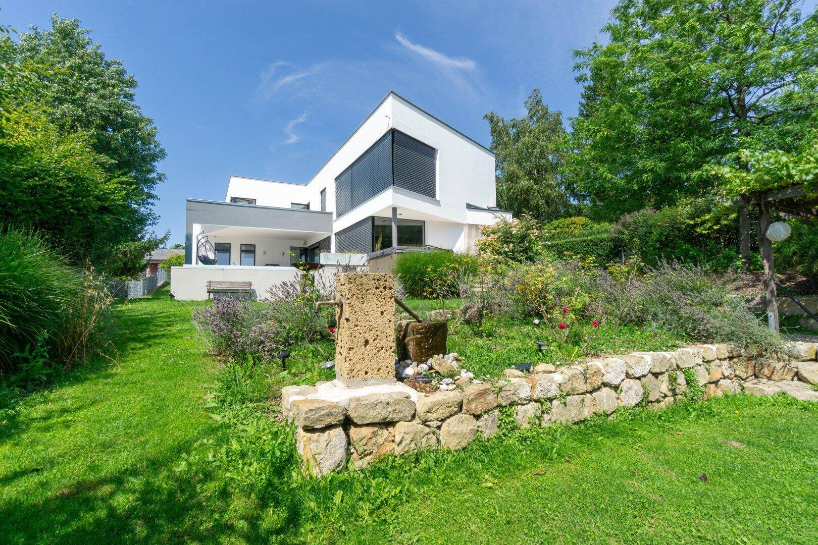 Gepflegter Gartenbereich mit Natursteinmauer, Zierbrunnen und Blick auf die moderne Villa.