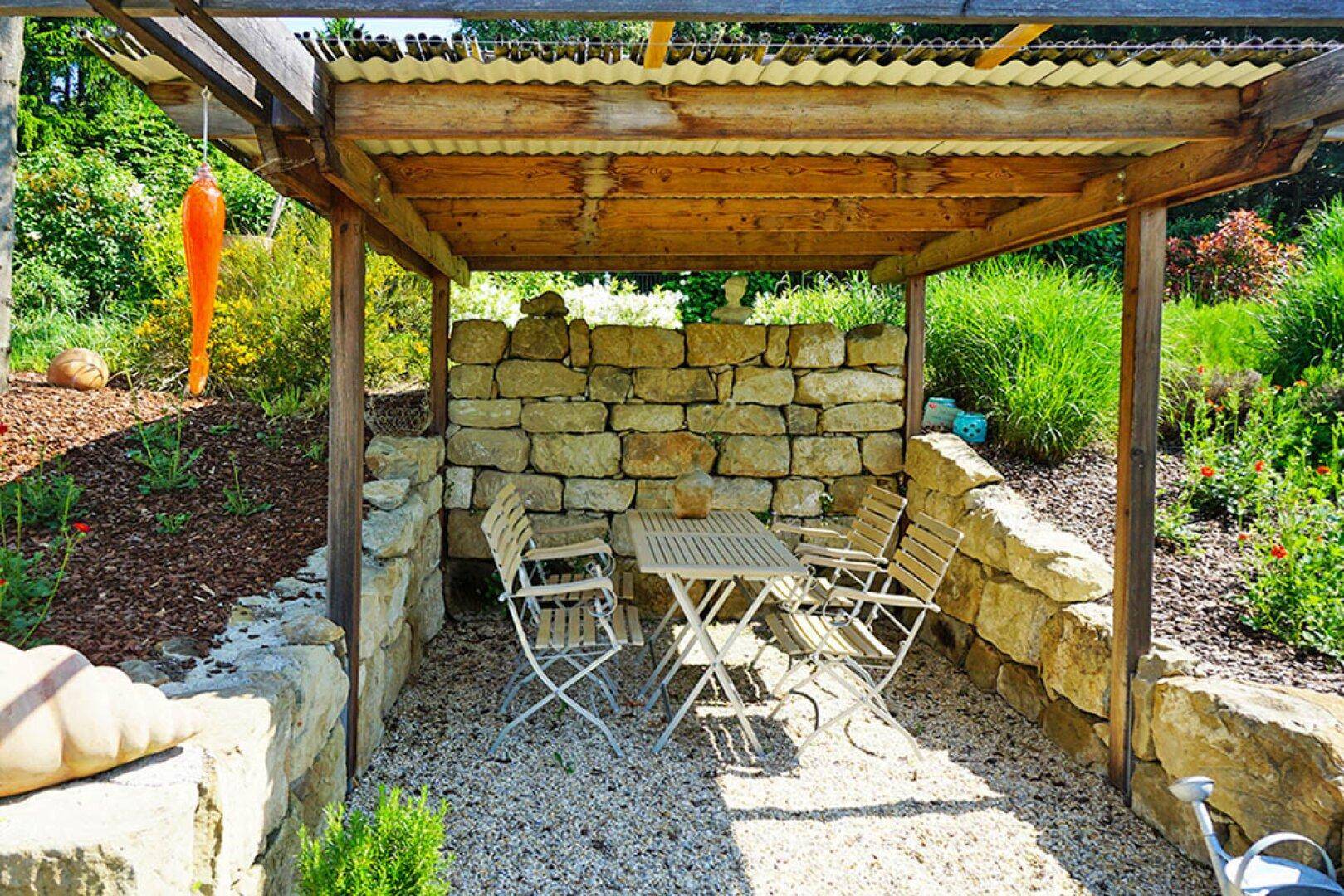 Gemütlicher Sitzbereich im Garten unter einer Pergola, umgeben von Natursteinmauern und Pflanzen.