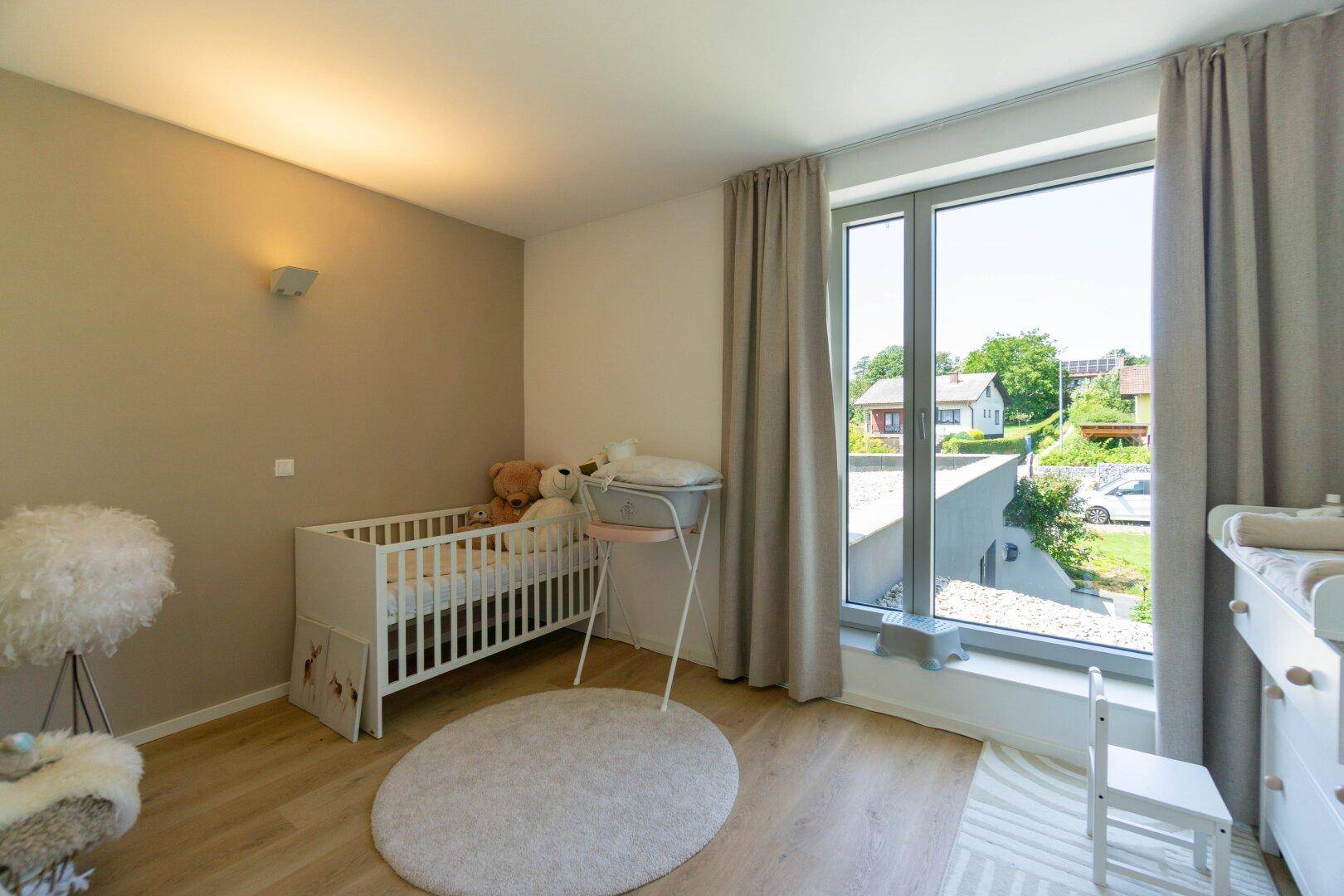 Helles Kinderzimmer mit Babybett, Wickeltisch und großem Fenster mit Blick ins Grüne.