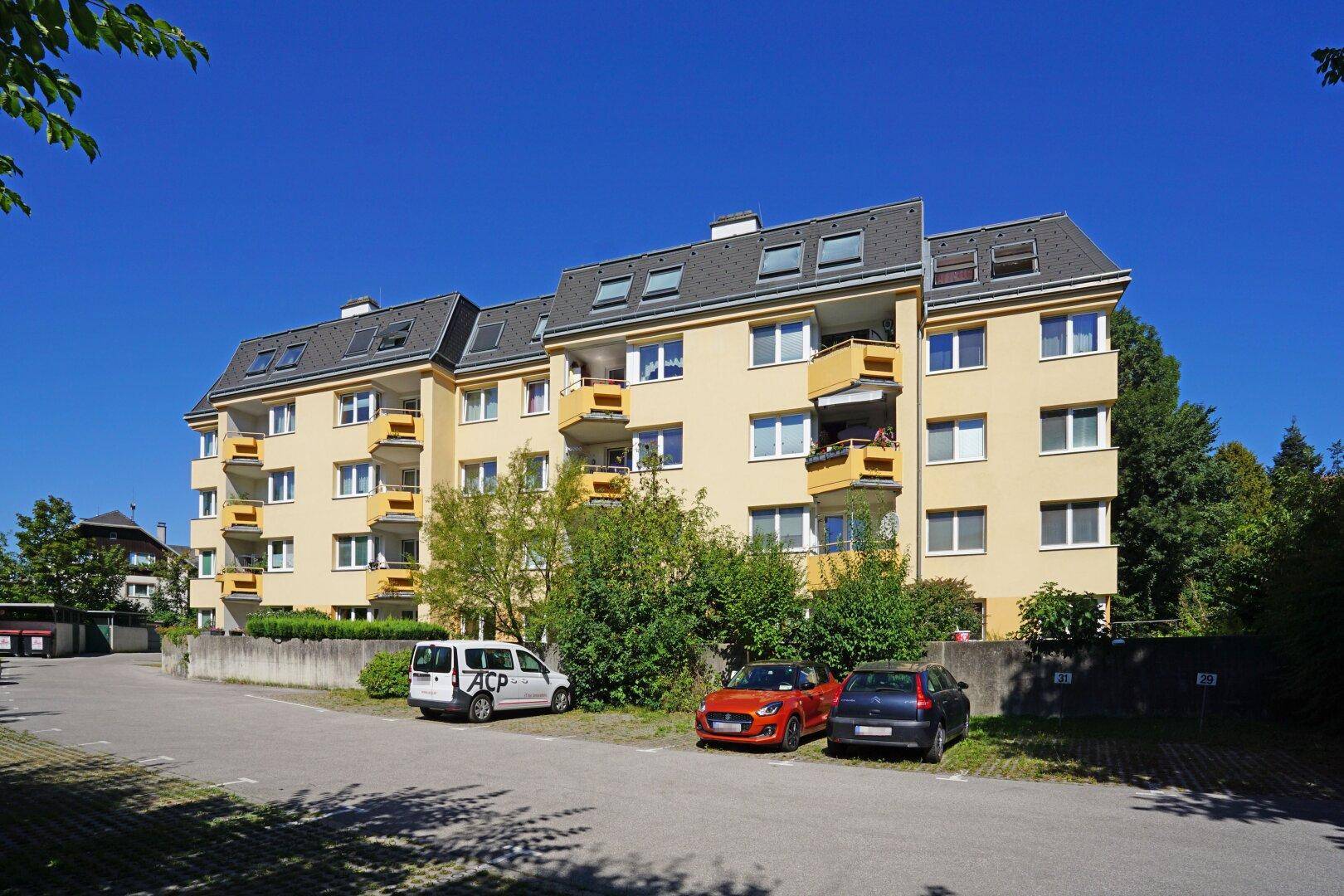 Mehrfamilienhaus mit gelber Fassade, Balkonen und Schrägdach in grüner Umgebung.