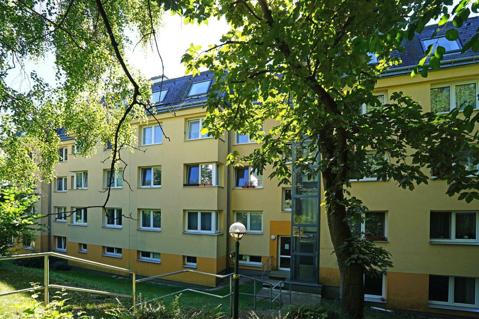Gelbe Wohnhausfassade mit vielen Fenstern und einem externen Aufzug im Grünen.