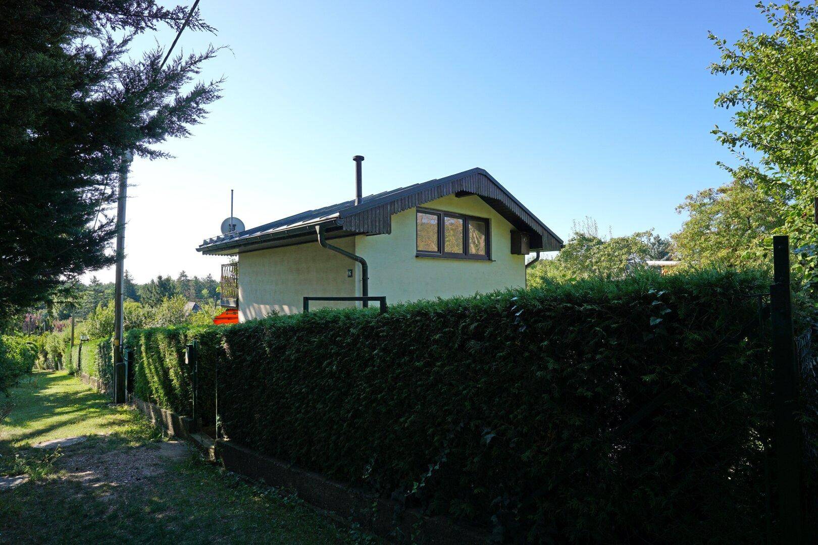 Kleines Haus mit Satteldach und grüner Hecke im Vordergrund unter blauem Himmel.