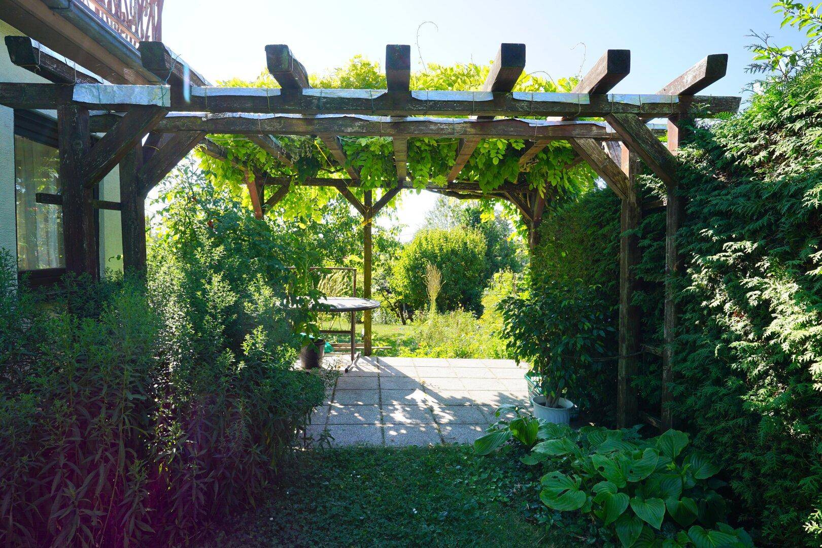 Steinplattenweg unter einer bewachsenen Holzpergola, die zu einem weiteren Gartenbereich führt.