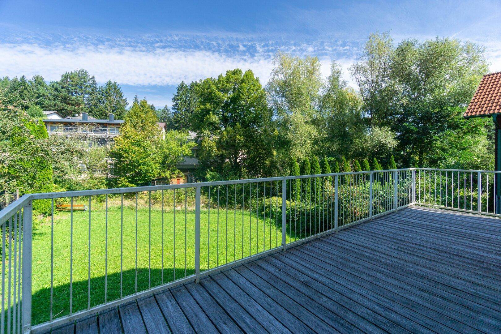 Großer Holzbalkon mit Metallgeländer und weitem Blick ins Grüne unter blauem Himmel.