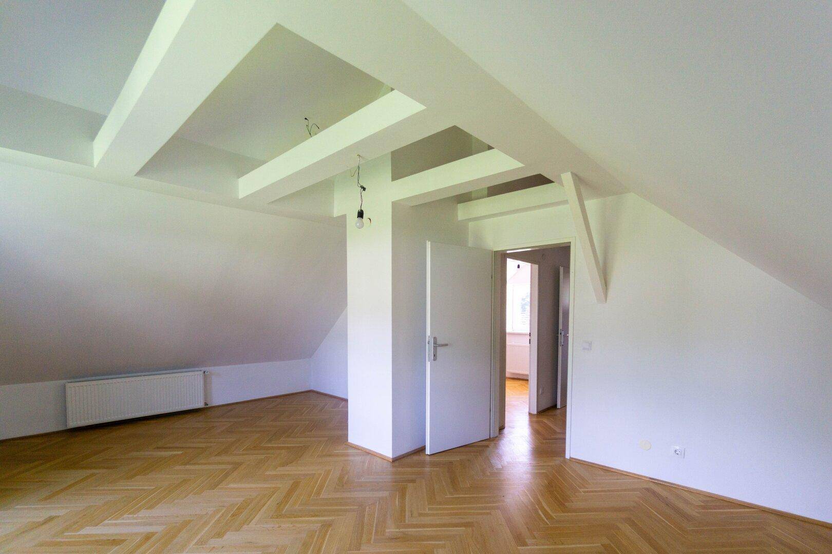 Dachgeschosszimmer mit sichtbaren Deckenbalken und hellem Parkettboden, unmöbliert und offen.