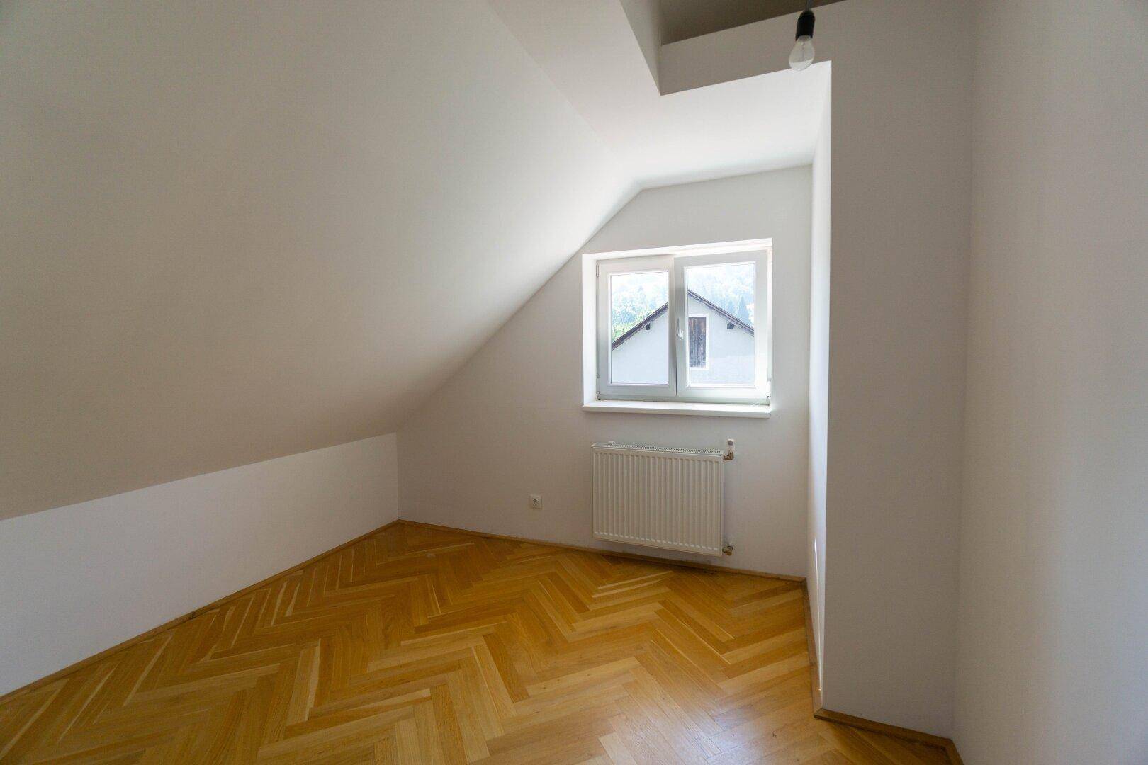 Helles Zimmer im Dachgeschoss mit schrägen Wänden, Fenster und Parkettboden.