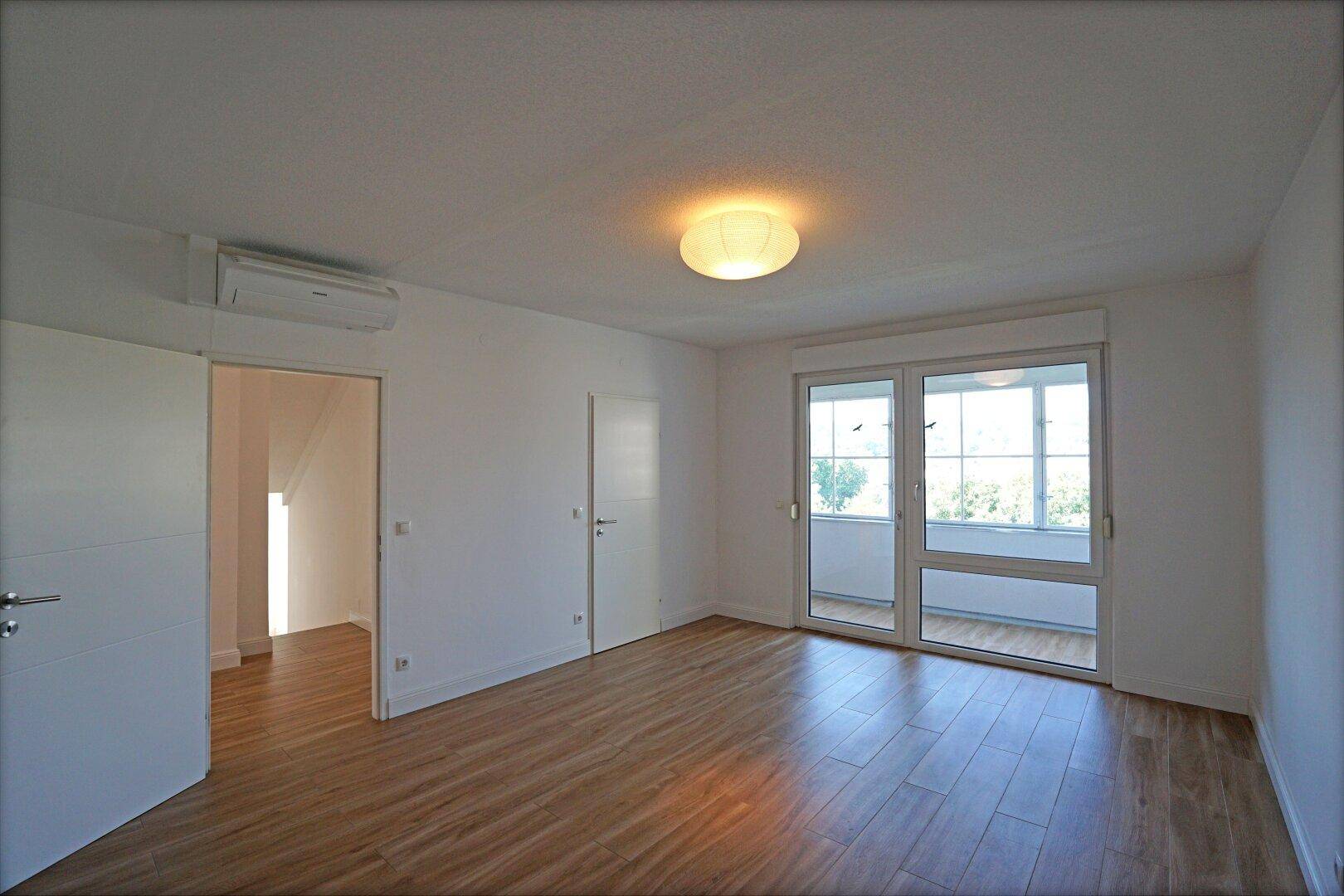 Helles Schlafzimmer mit Holzboden, großen Fenstern und Zugang zu einem Balkon.