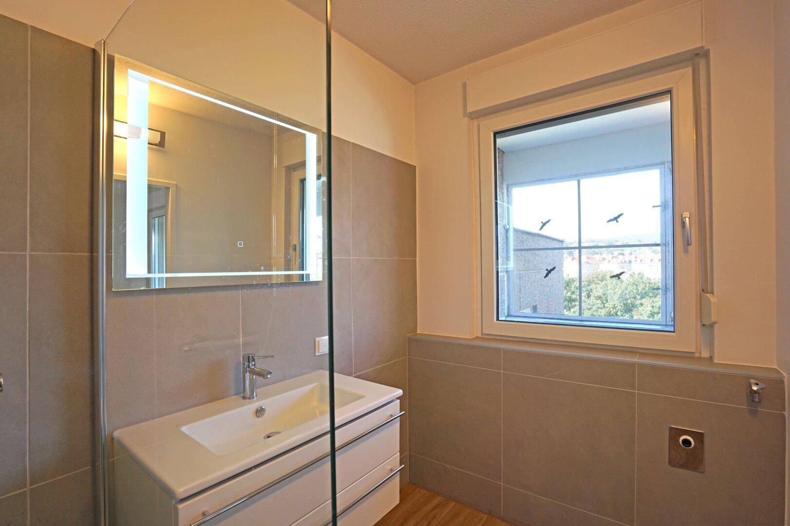 Modernes Badezimmer mit Waschtisch, beleuchtetem Spiegel und Fenster für Tageslicht.