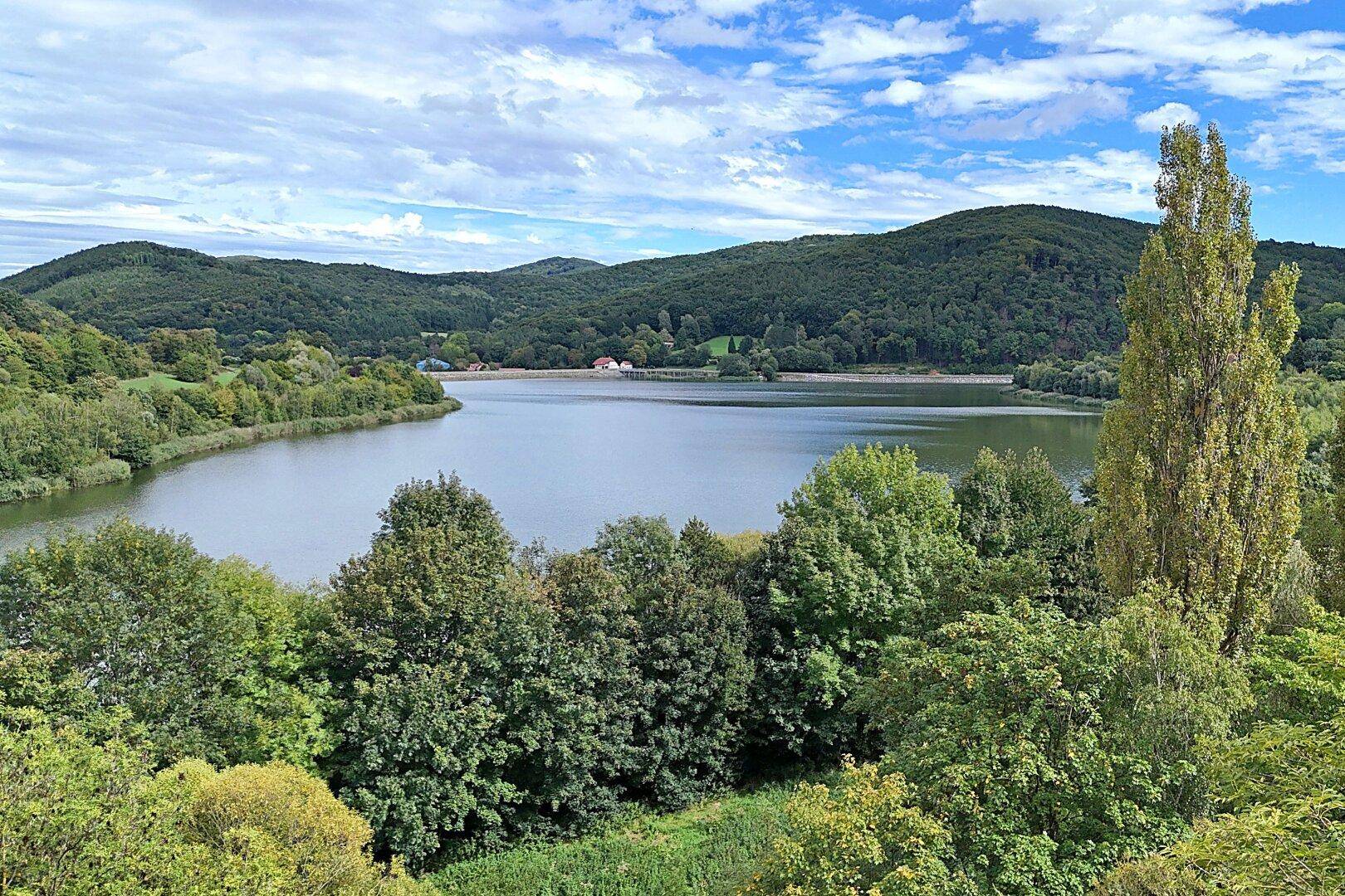 Malerischer Blick auf einen ruhigen See, umgeben von bewaldeten Hügeln unter einem blauen Himmel.