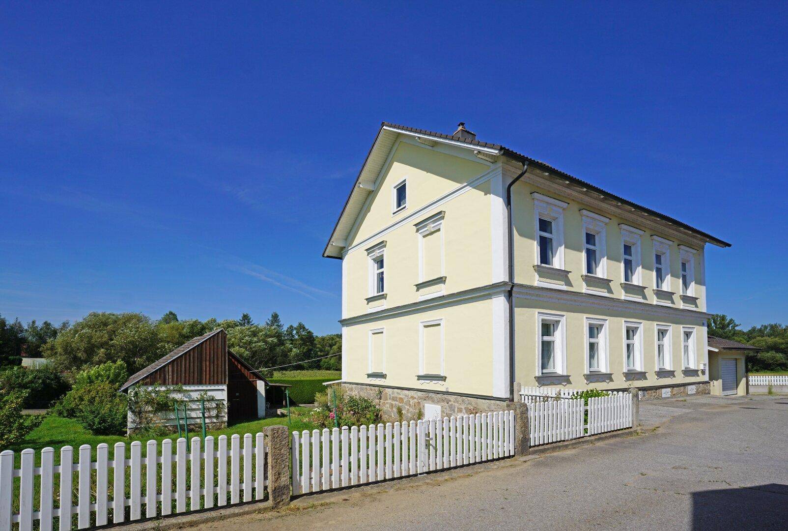 Helles, freistehendes Haus mit gelber Fassade, weißem Zaun und ländlicher Umgebung unter blauem Himmel.
