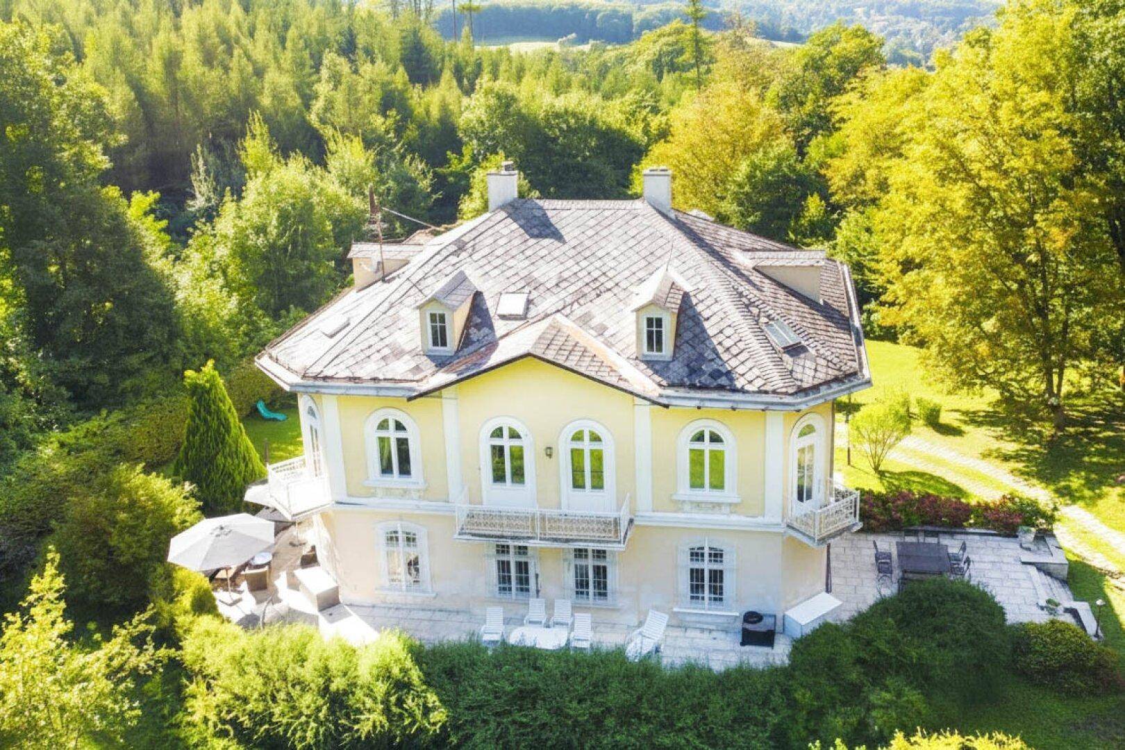 Luftaufnahme einer historischen Villa mit Mansarddach, umgeben von üppigem Grün und Wald.