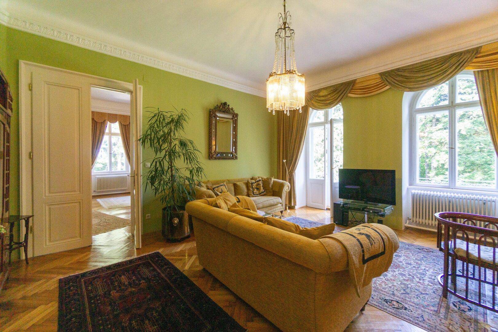 Helles Wohnzimmer mit grünen Wänden, Parkettboden und eleganten Vorhängen, mit Blick ins Grüne.