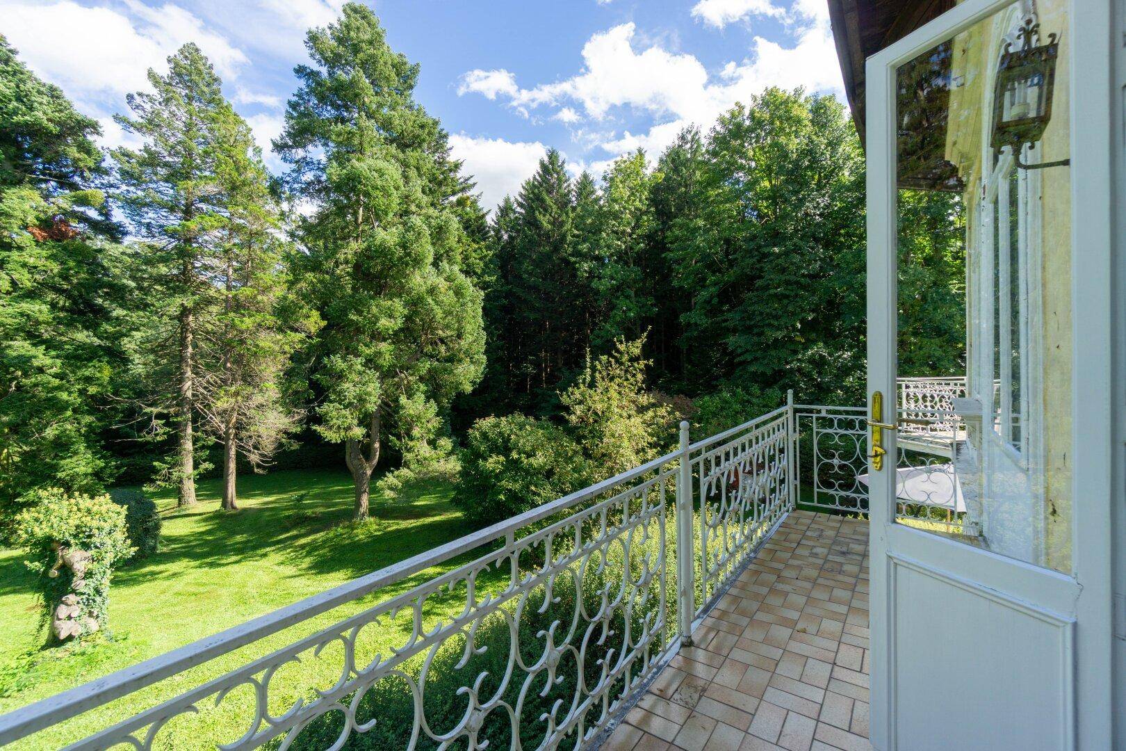 Balkon mit schmiedeeisernem Geländer und weitem Blick auf den grünen Garten und Wald.