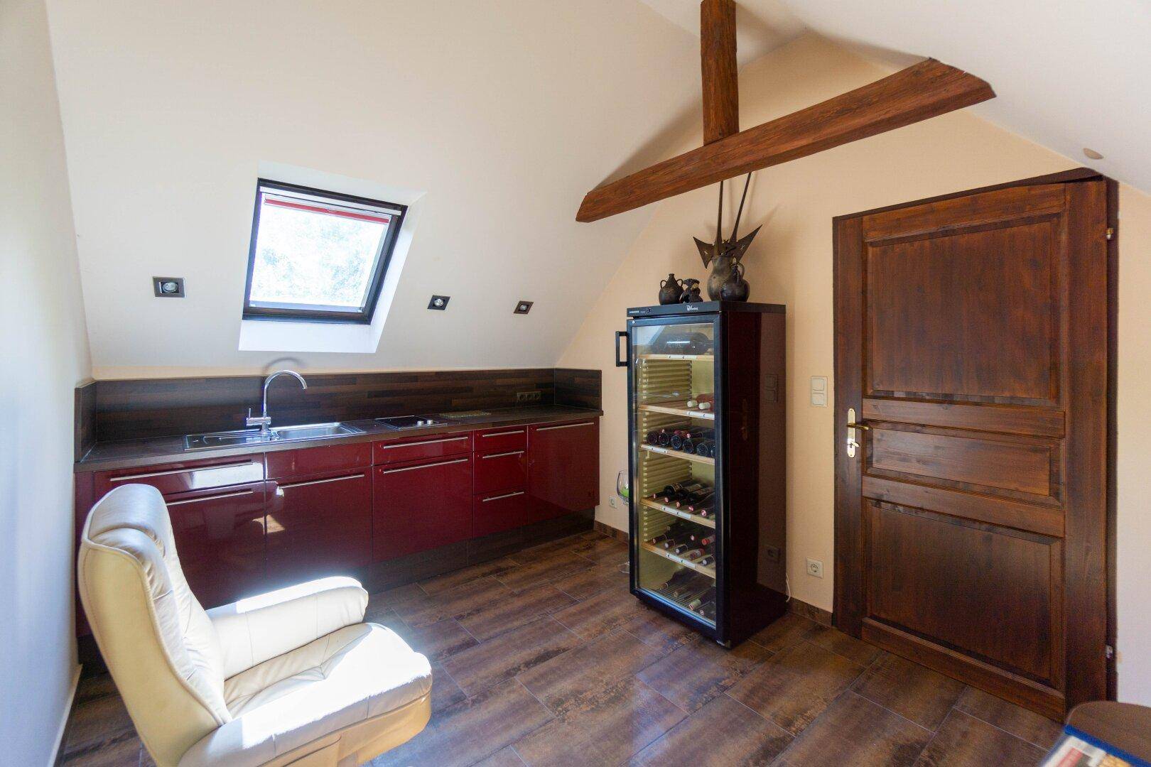 Dachbodenraum mit kleiner Küchenzeile, Weinkühler und Holzbalken unter schräger Decke.