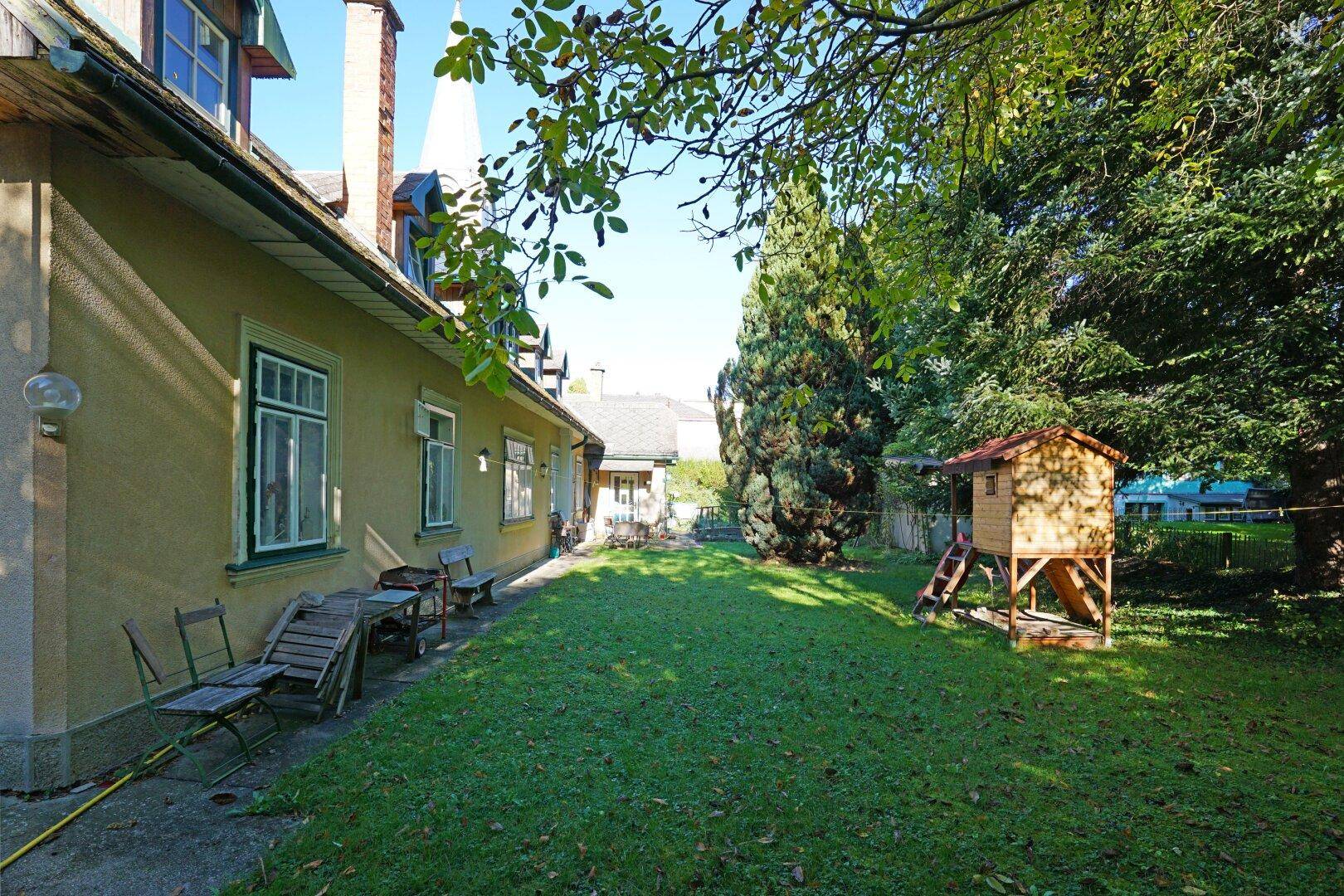 Großer Gartenbereich neben dem Haus mit Rasenfläche, Bäumen und einem kleinen Spielhaus für Kinder.