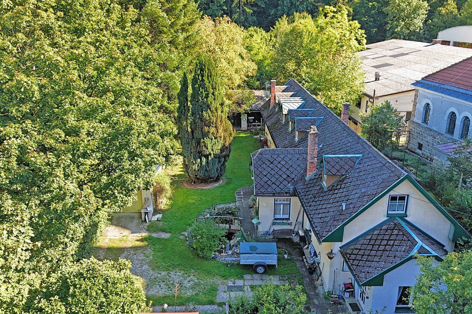 Luftaufnahme des Anwesens mit dem Haus, dem Garten und umliegenden Bäumen unter blauem Himmel.