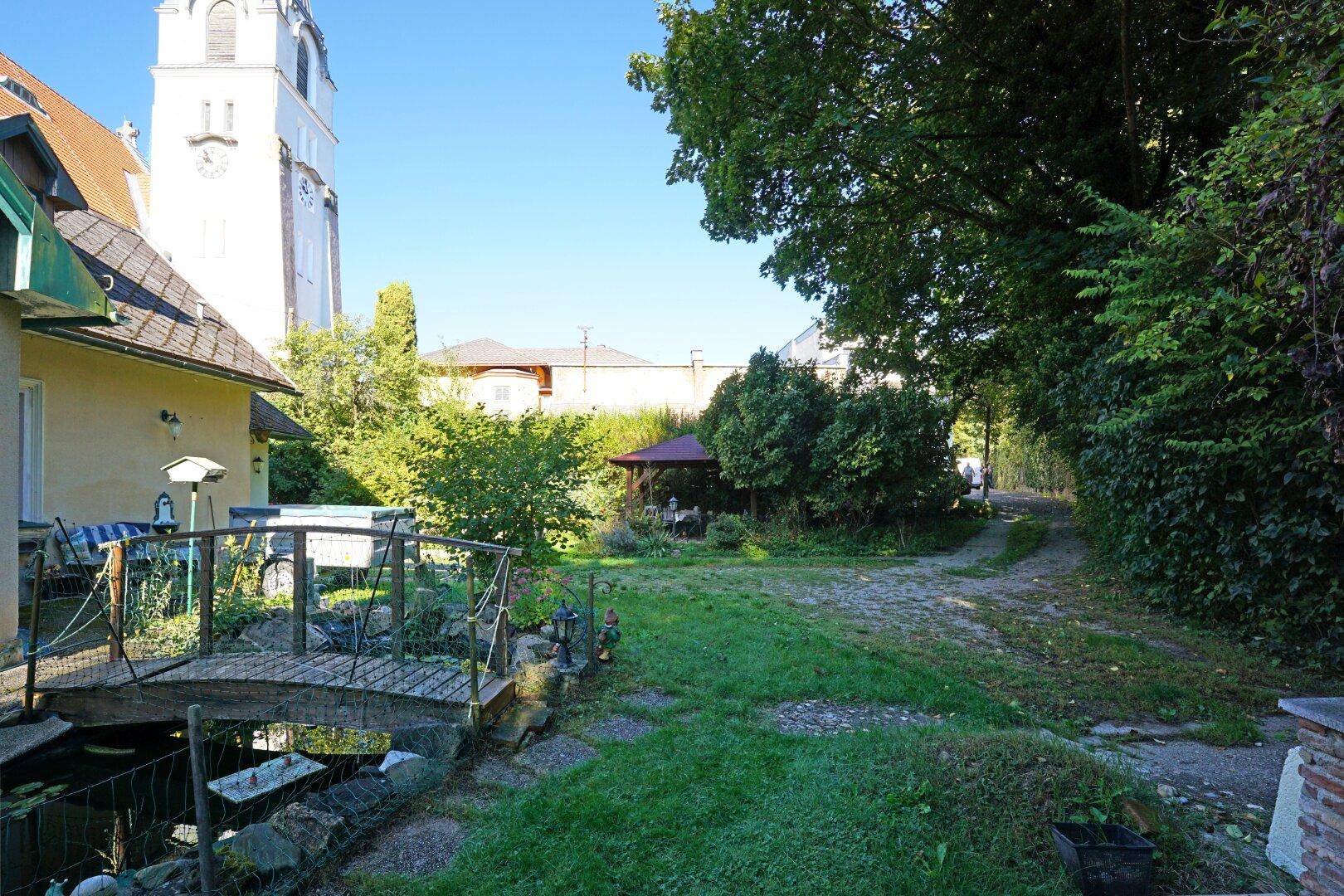 Idyllischer Gartenbereich mit kleinem Teich, Holzbrücke und einem Pavillon im Hintergrund.