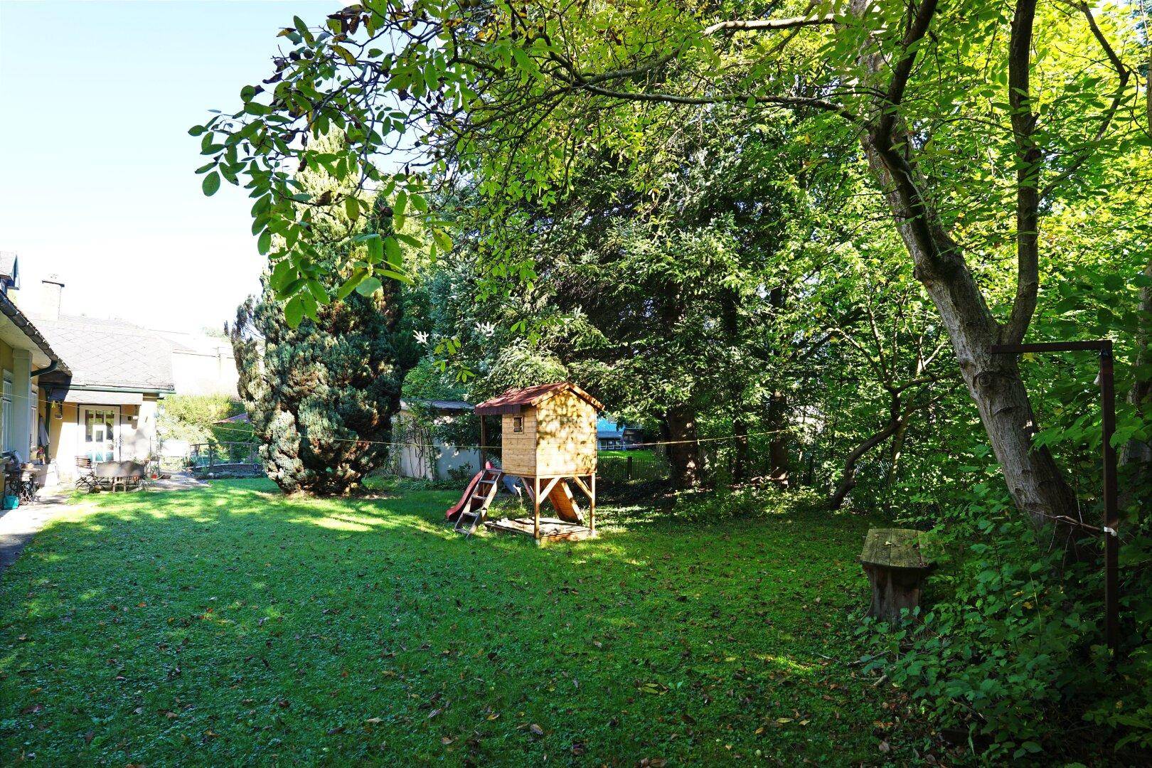 Weitläufiger Garten mit Rasen, altem Baumbestand und einem Holzspielhaus für die Kleinen.