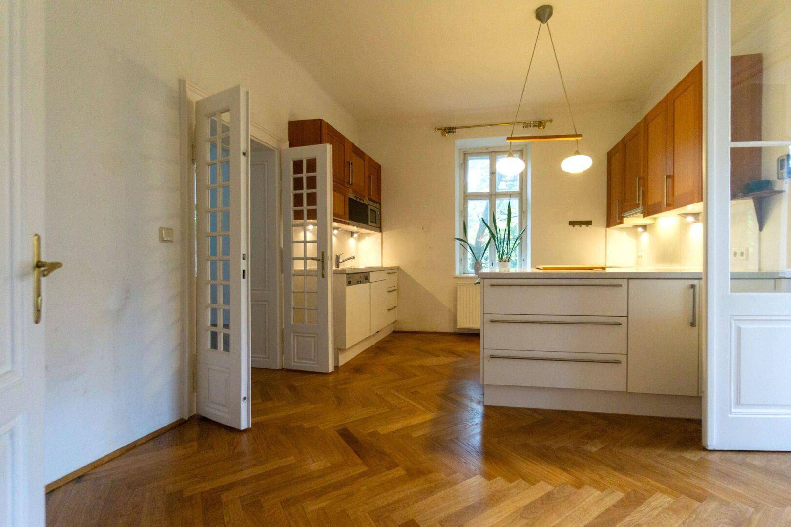 Helle Küche mit Fischgrätparkett, weißen Schränken und Zugang zu weiteren Räumen.