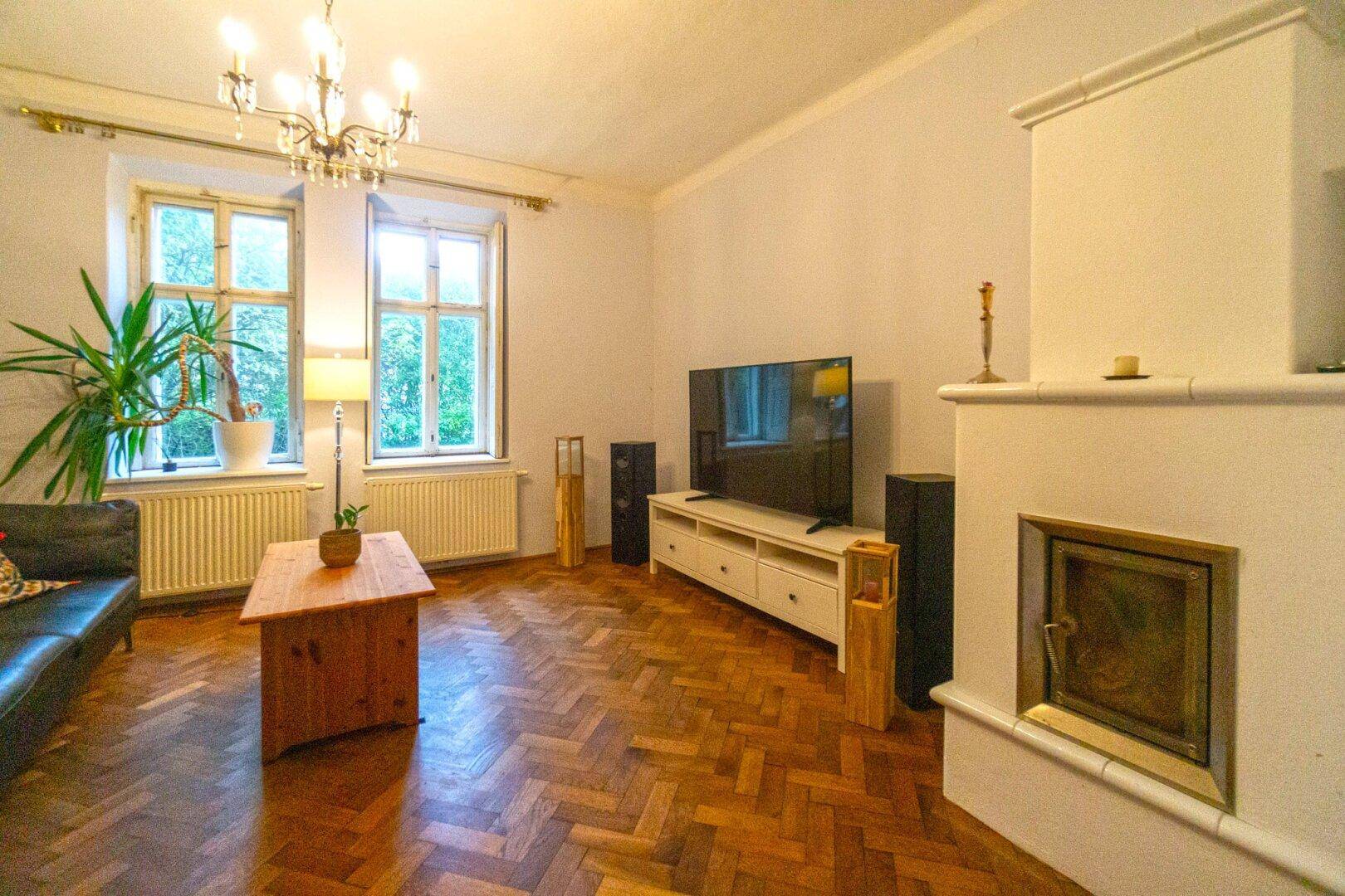 Helles Wohnzimmer mit Kaminofen, Fernseher und klassischem Fischgrätparkett.