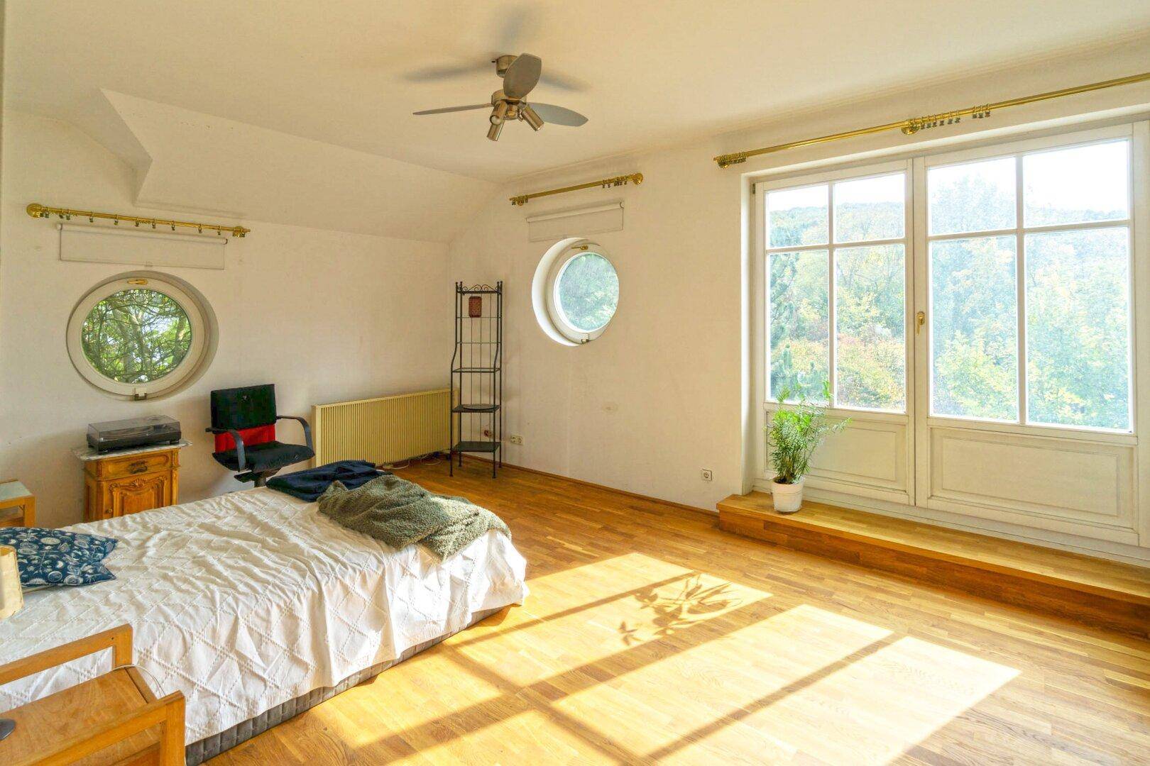 Sonniges Schlafzimmer mit Holzboden, runden Fenstern und Blick ins Grüne.