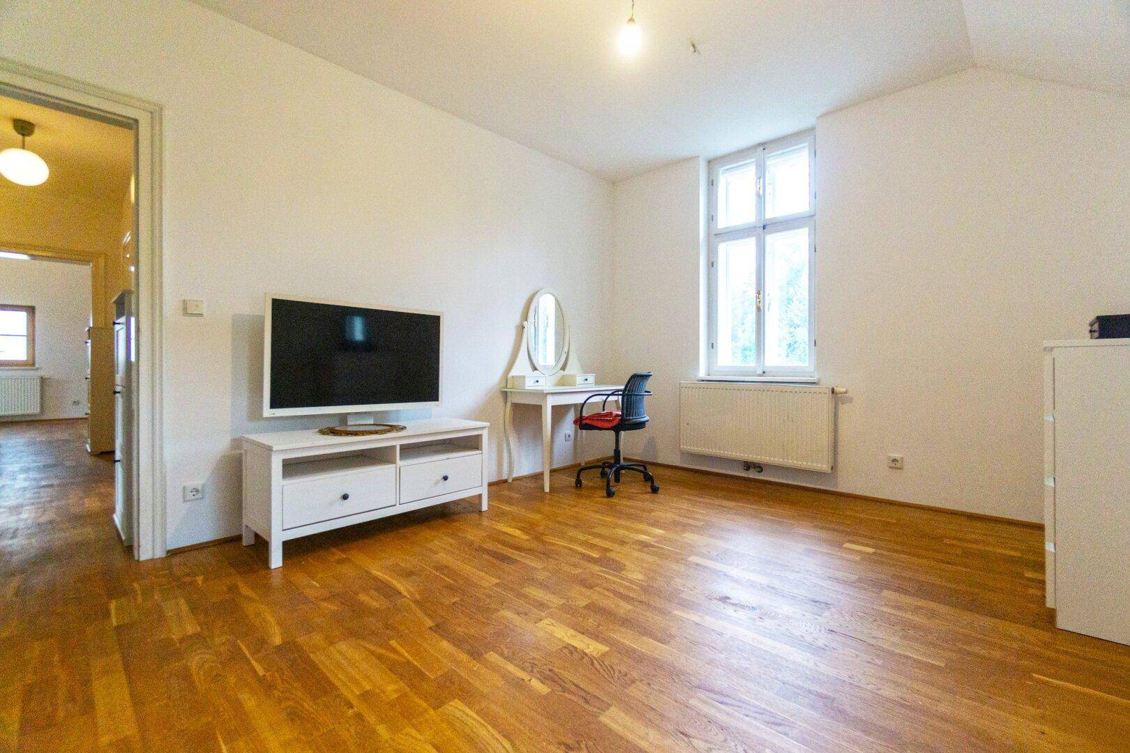 Geräumiges Zimmer mit Holzboden, großem Fenster und einfacher Einrichtung.
